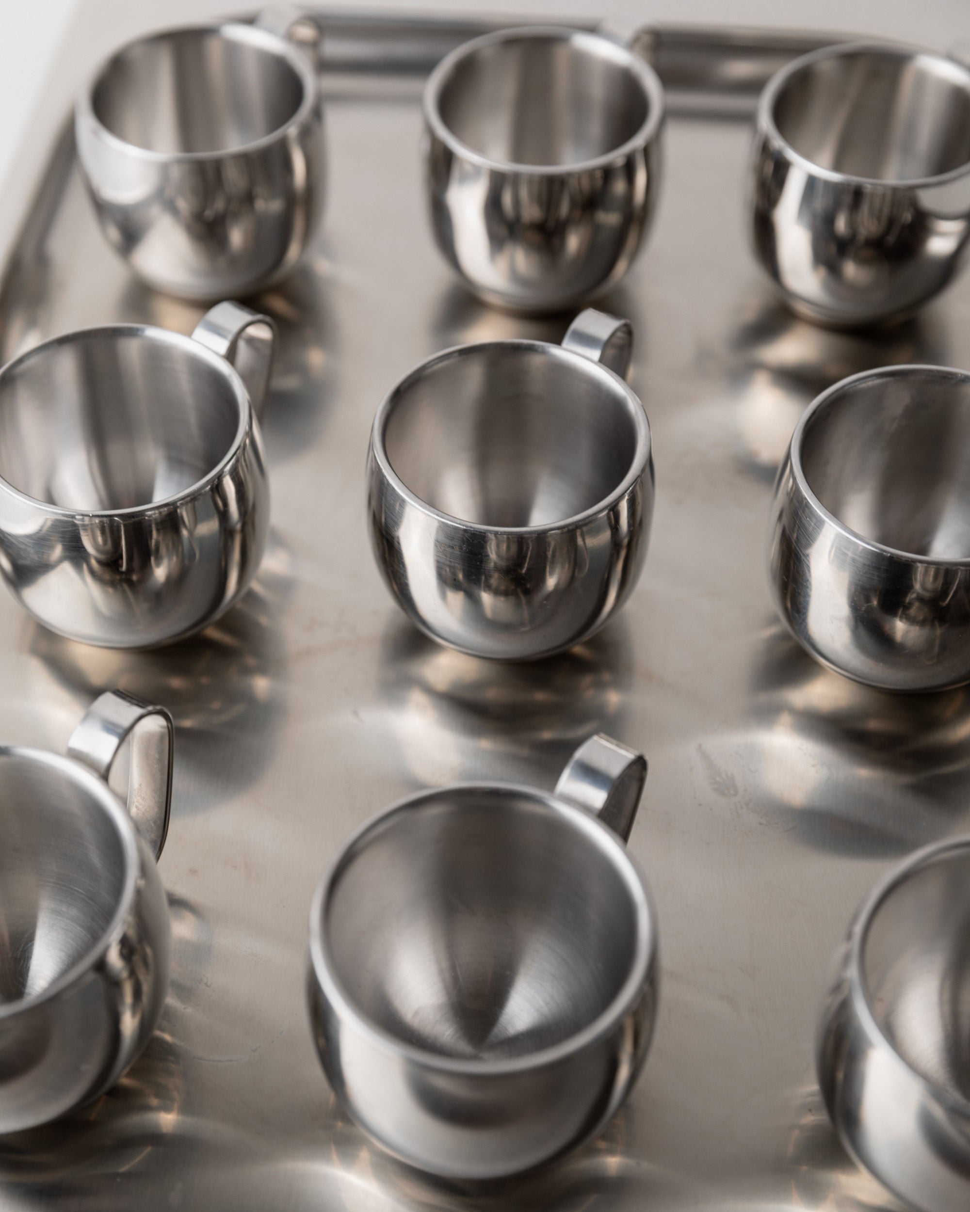 A close-up of nine stainless steel Coffee set by IMCO 70s cups from Bottega Jacobs, arranged in a grid on a reflective metal surface. This minimalist design showcases shiny, uniform mugs, creating a symmetrical and organized appearance.