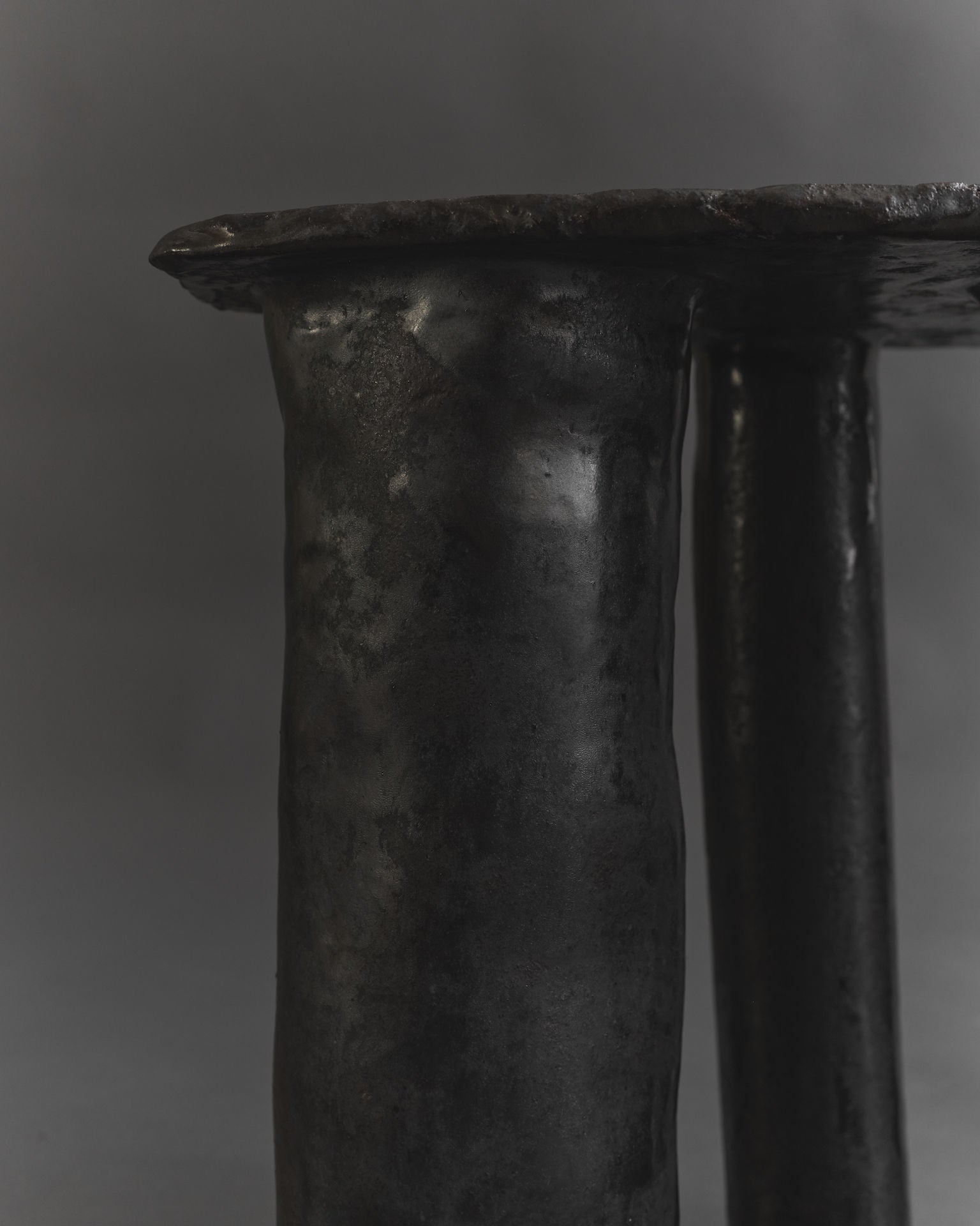 A close-up of the Cleo Ceramic Side Table by Ombia Studio showcases a dark, textured metal sculpture with two cylindrical columns supporting a flat, round top. The hand-crafted surface has a rugged, slightly uneven texture reminiscent of black glaze against a smooth gray background.