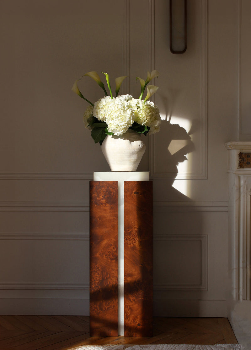 A sunlit room features the Marbera 'Como' Plinth in white onyx and brown wood, topped with a ceramic vase of white flowers. Soft shadows fall on a white wall with molding, while a brown wood fireplace peeks in from the right.