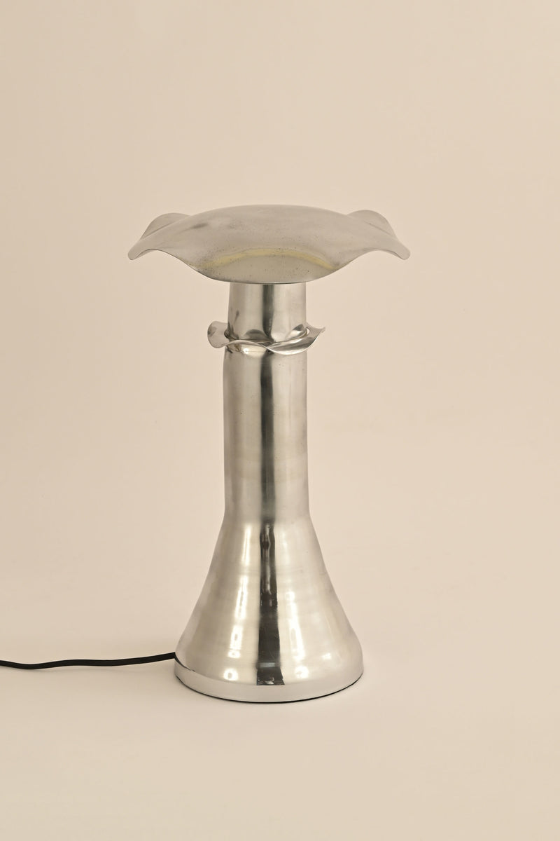 The LMNOH Funghi Table Lamp features a modern metallic design with a flared base and a flower-shaped translucent top, casting ambient light. Its black power cord accentuates the minimalist look against a plain light background.