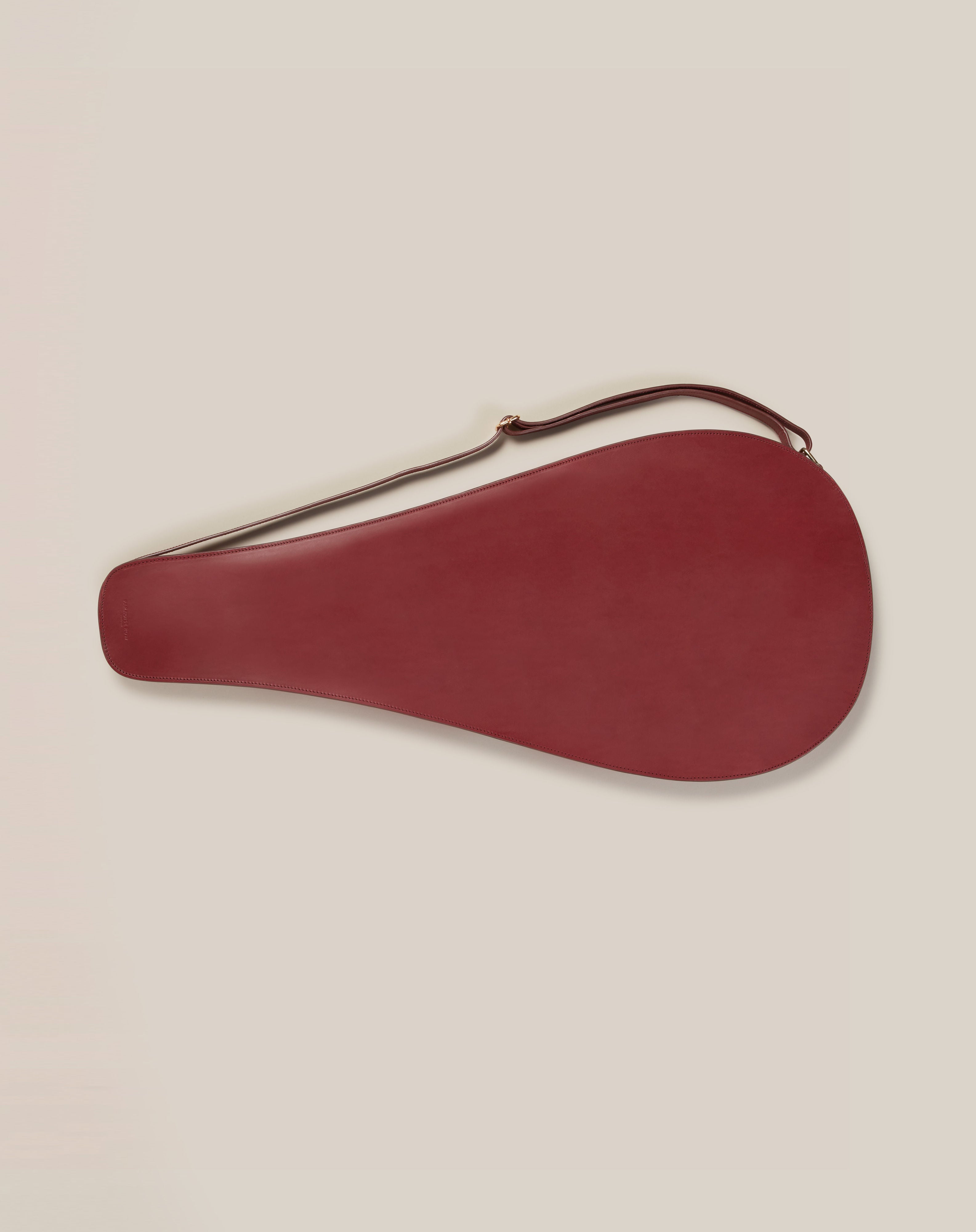 A red, teardrop-shaped Leather Tennis Racket Cover with a brown strap, crafted from sustainable vegetable-tanned leather by Paradise Row, lies against a light beige background.