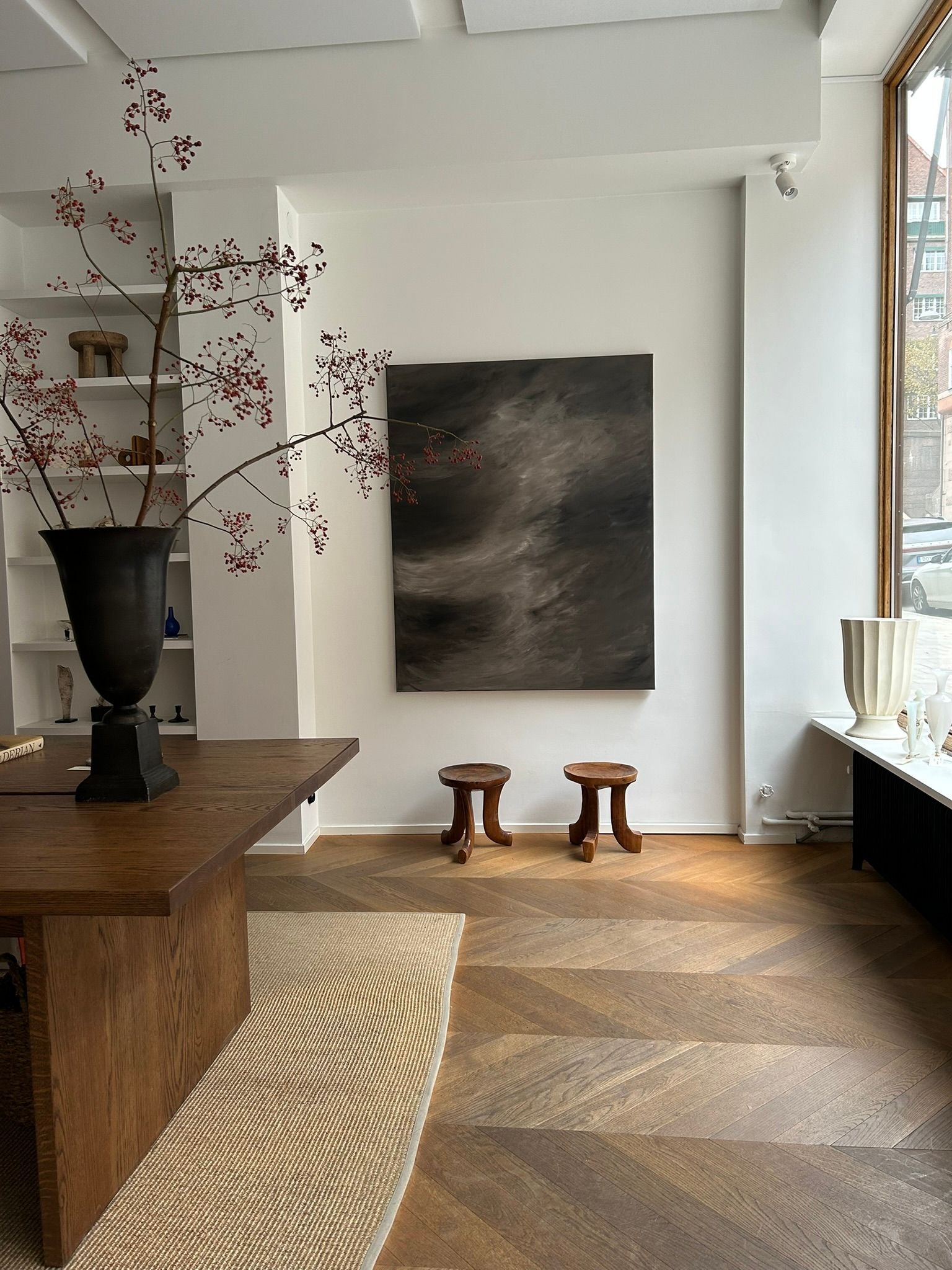 A modern room is adorned with a large wooden table, alongside a tall vase filled with branches and two small stools. On the wall, Mattias Storm's dark abstract painting titled "Breath of Dawn" adds an artistic touch. Natural light floods through a tall window on the right, beautifully highlighting the wooden floor and mixed media elements throughout.