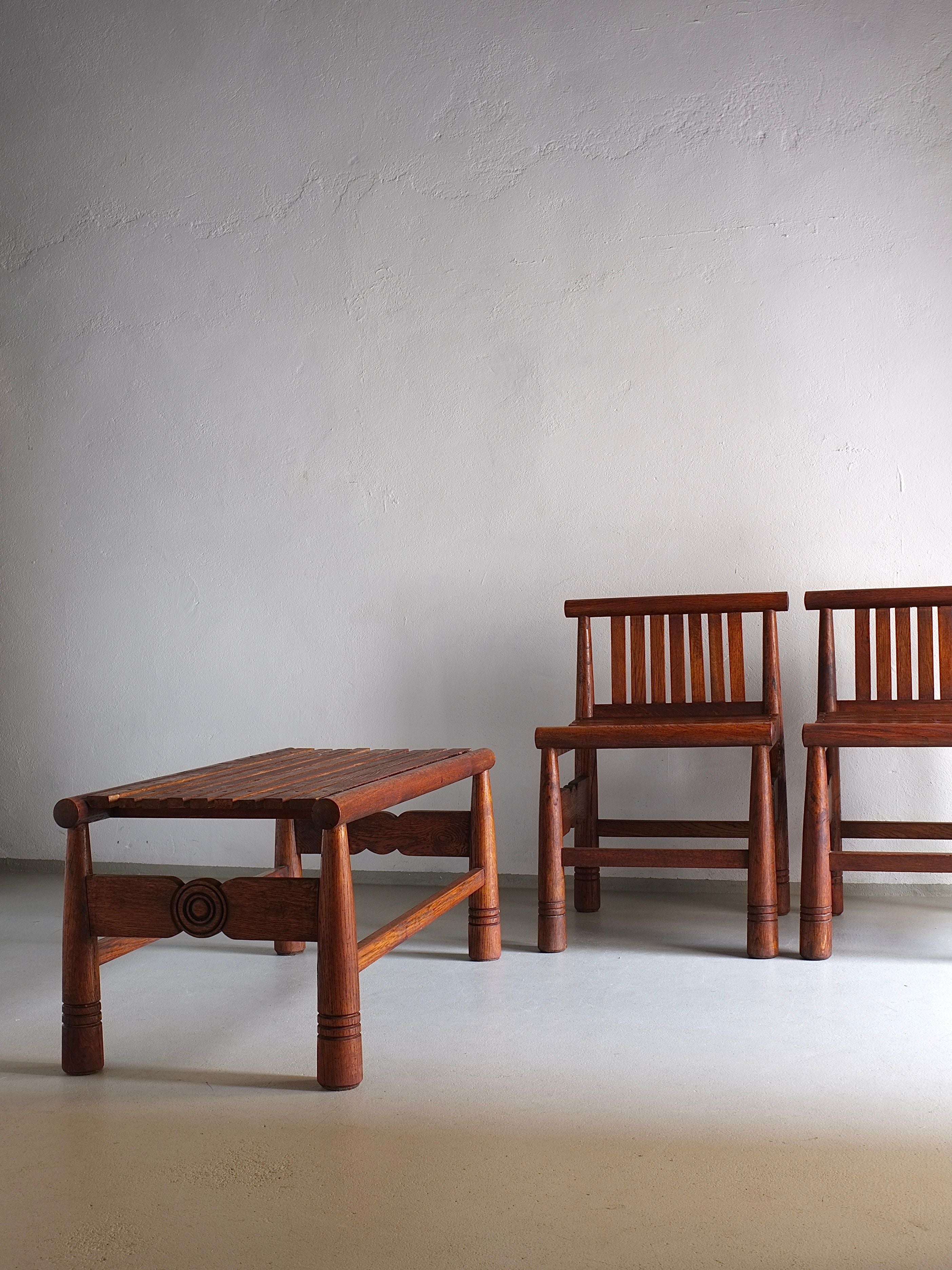 A simple and minimalistic room features two wooden chairs with vertical slats and a Carved Dark Oak Bench from the 1960s, designed by Veter Vintage, showcasing intricate circular details on the legs. The furniture is all arranged against a plain, light-colored wall. The vintage pieces exude a rustic and handcrafted appearance.