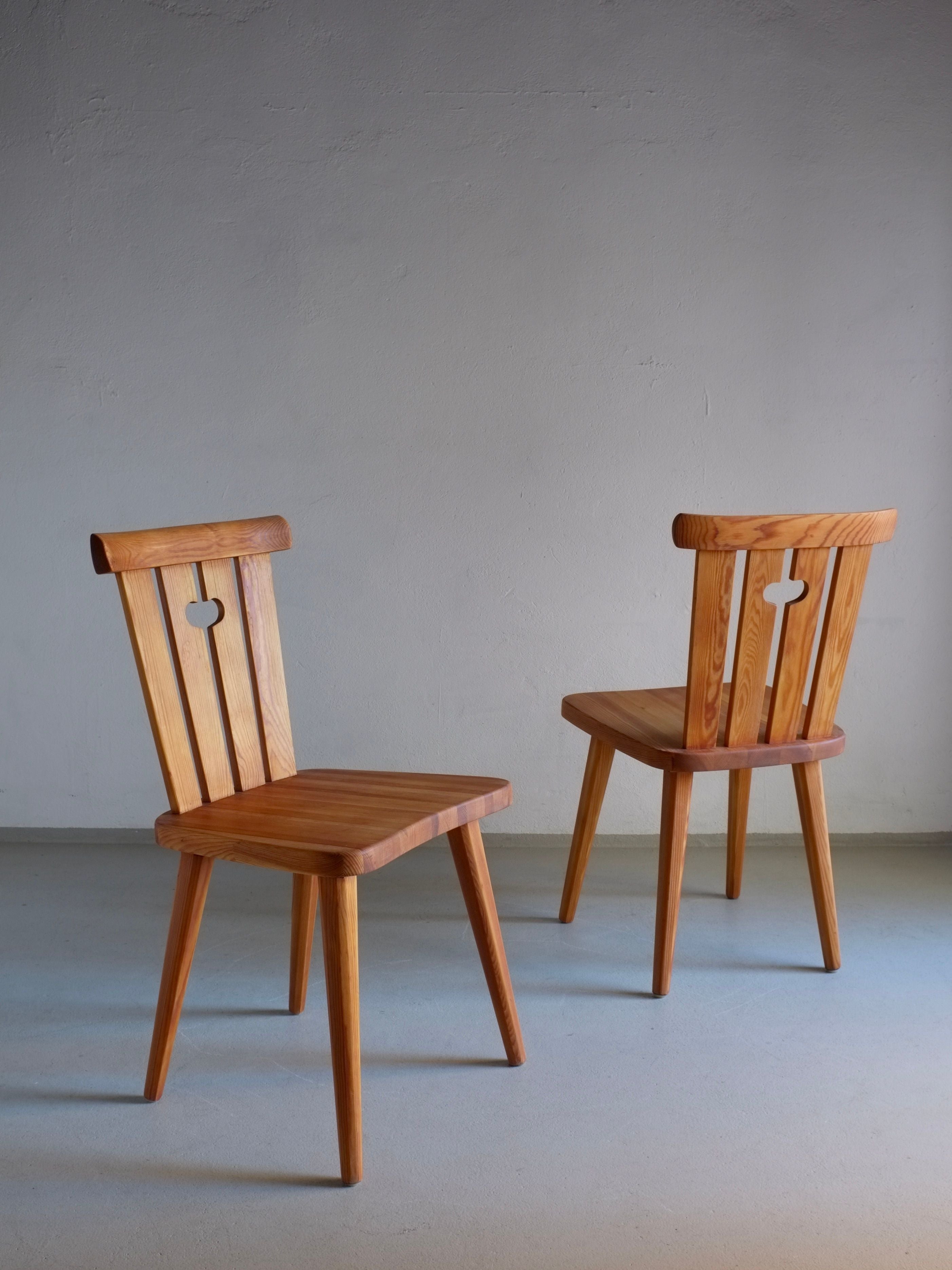 Two rustic pine chairs, expertly restored and oiled to perfection, feature heart-shaped cutouts on the backrest. Set against a simple light-colored floor, these creations by Göran Malmvall for Veter Vintage exude a natural wood finish that complements the minimalist light gray wall.