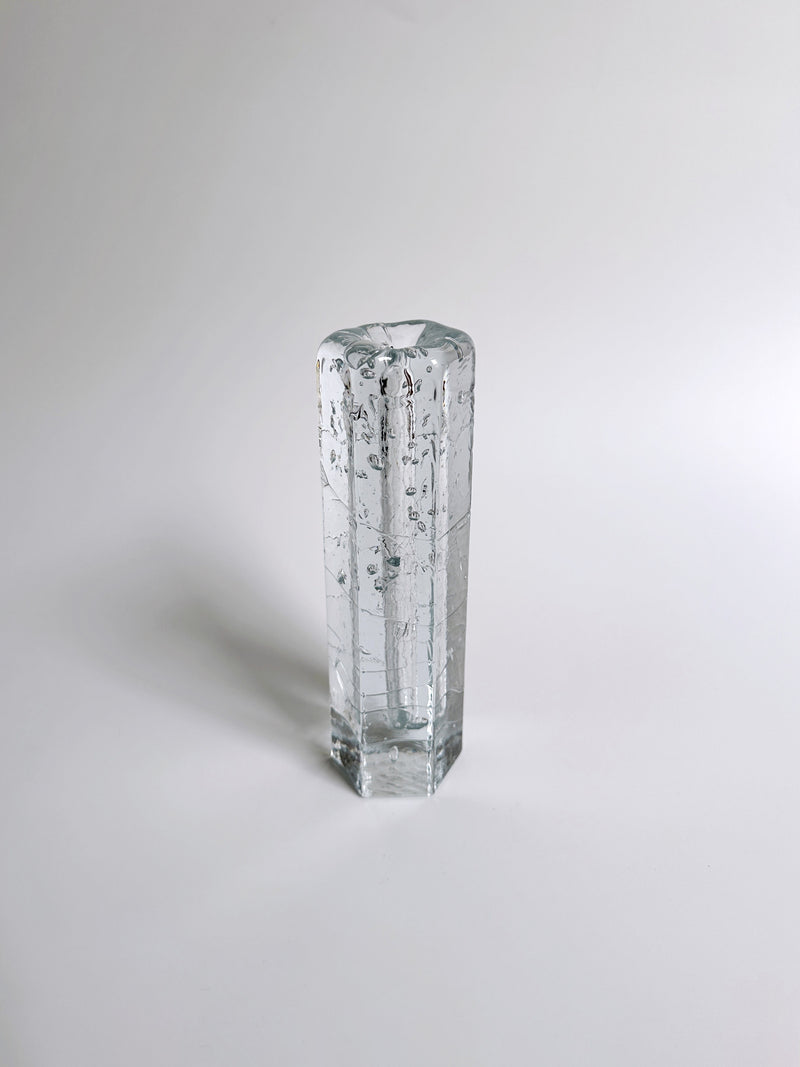 A tall, clear glass vase with an octagonal shape stands upright against a white background. This 70s Swedish glass vase by Dodo Vintage, crafted using the Ice glass technique, features small, dispersed bubbles within the glass, giving it a textured appearance. Its surface is smooth and subtly reflective.