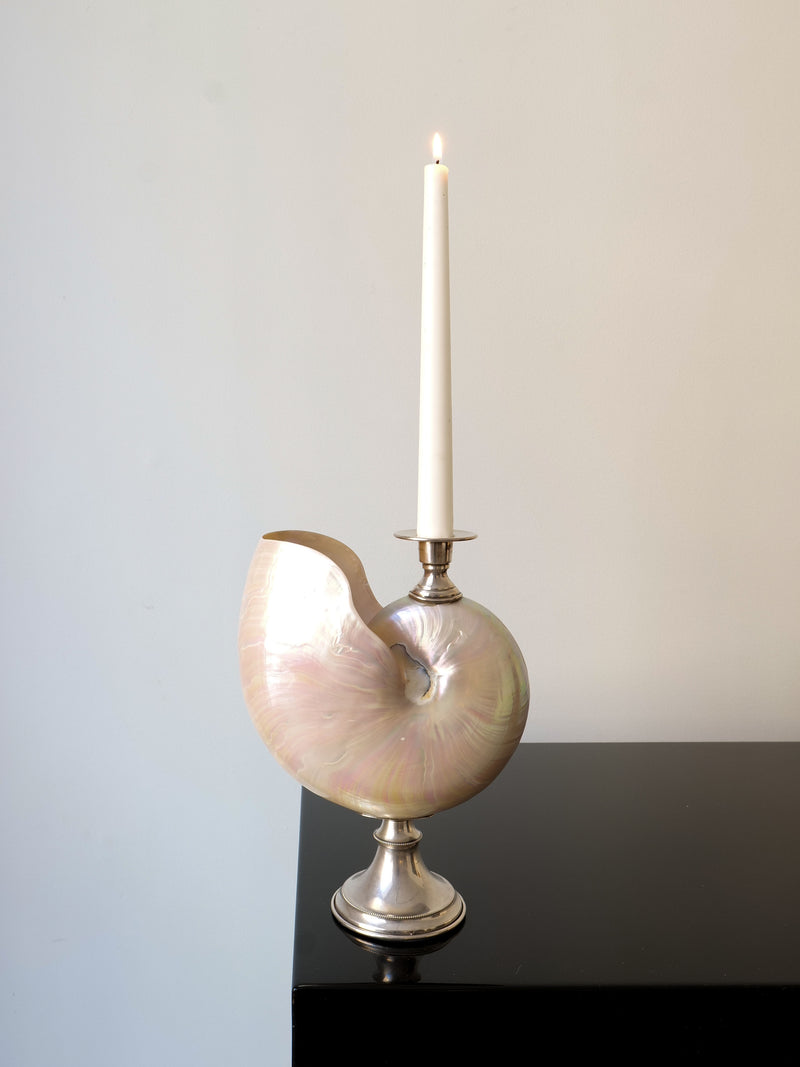 A white candle flickers in the Nautilus Silver Candle Holder by Collection apart, beautifully crafted from a nautilus shell. It rests elegantly on a shiny black surface against a plain light-colored background, exuding sophistication.