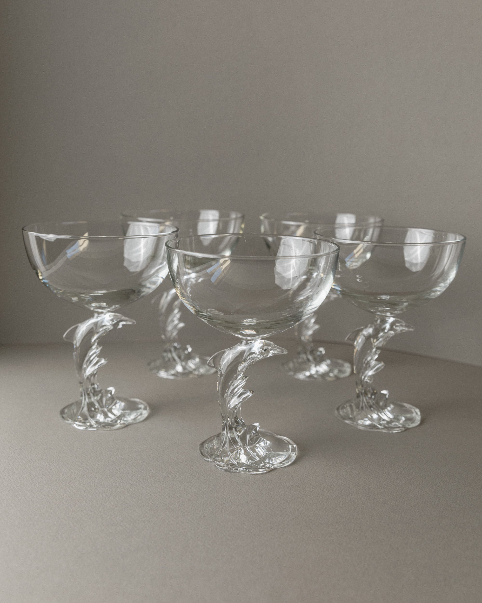 Five Bottega Jacobs Luminarc Delfin Champagne Coupes from the 80s, featuring intricately twisted stems, are symmetrically arranged on a neutral surface against a soft gray background.
