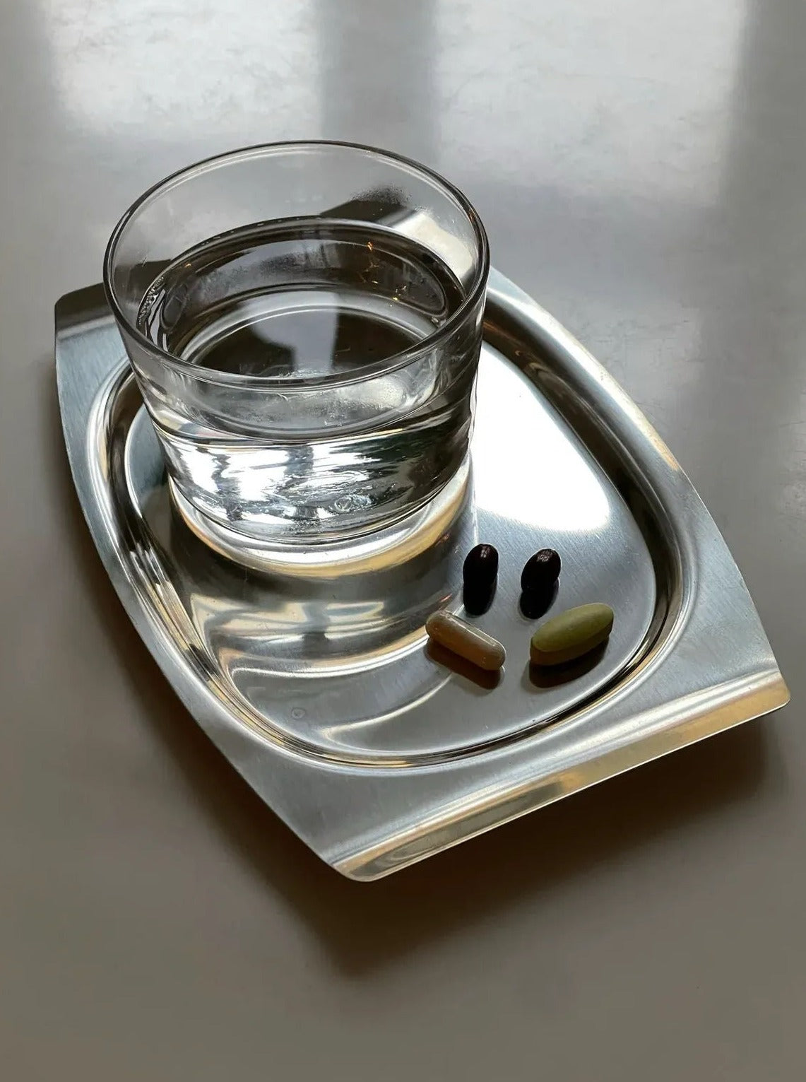 A transparent glass of water on a Collect Cph stainless steel tray, accompanied by four capsules in varying colors, resting on a matte gray surface.