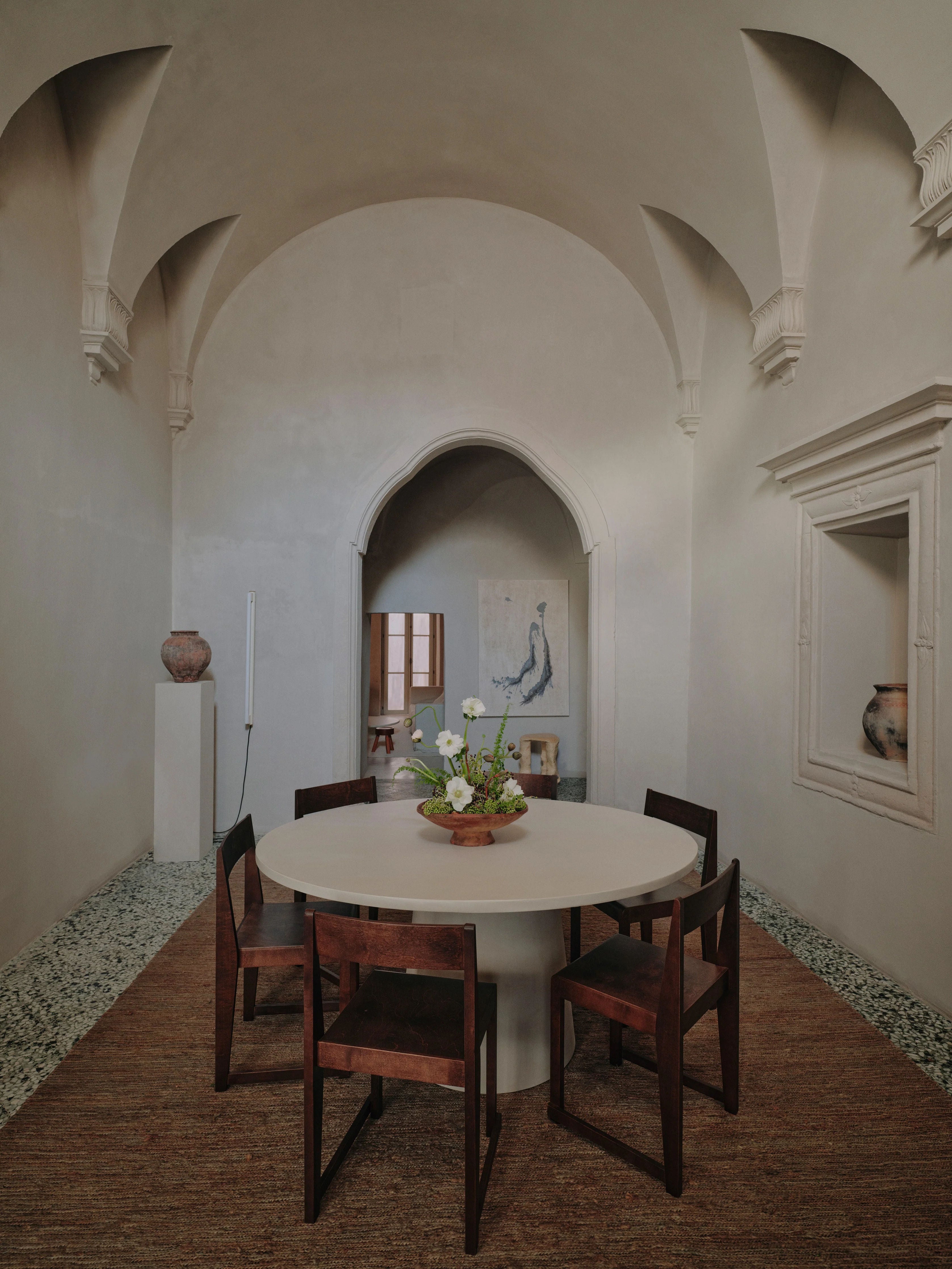 A minimalist dining area highlights the "Core" Dining Table by MarlotBaus, featuring a scratch-resistant microcement round tabletop. Accompanied by four wooden chairs placed on a textured rug, the room showcases arched architecture and neutral-toned walls with ornate ceiling details. A vase with flowers sits at the center of the table, enhanced by modern art and pottery decor.