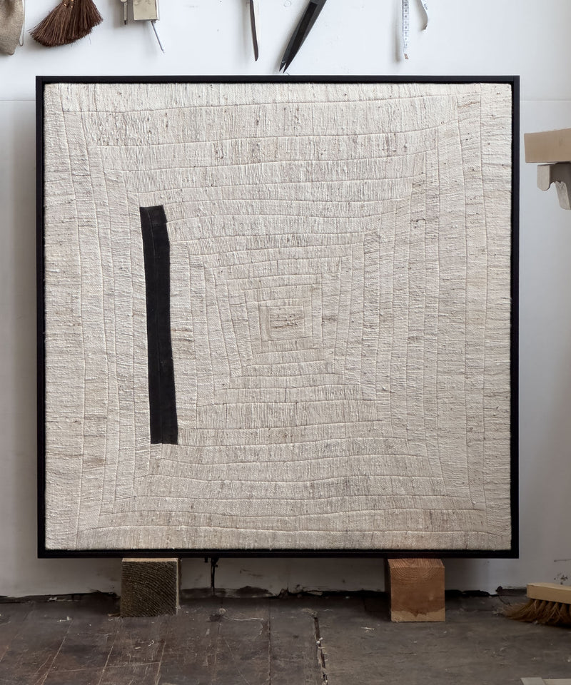 The "to tilt, 2024" artwork by Bec Kirby is a square piece crafted from handwoven kapok fiber cloth. It showcases textured, concentric hand-stitched lines forming a spiral effect on cream-colored fabric, with a black vertical stripe along the left side. This unique artwork is displayed against a white wall and rests on solid oak blocks.