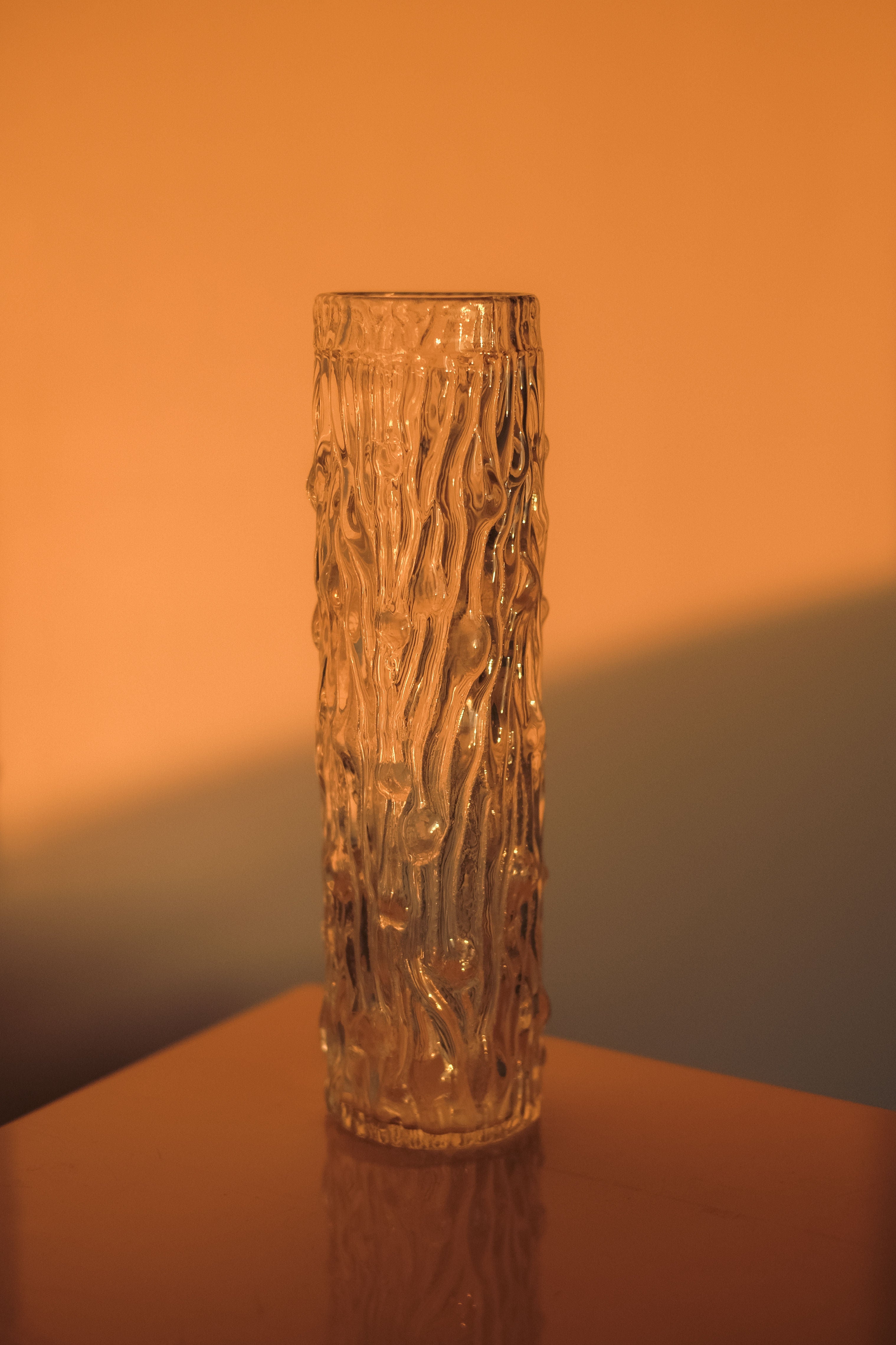 A Small Glass Cilinder Vase by septembre studios, featuring a textured design, stands on a reflective surface under warm orange lighting that casts soft shadows.