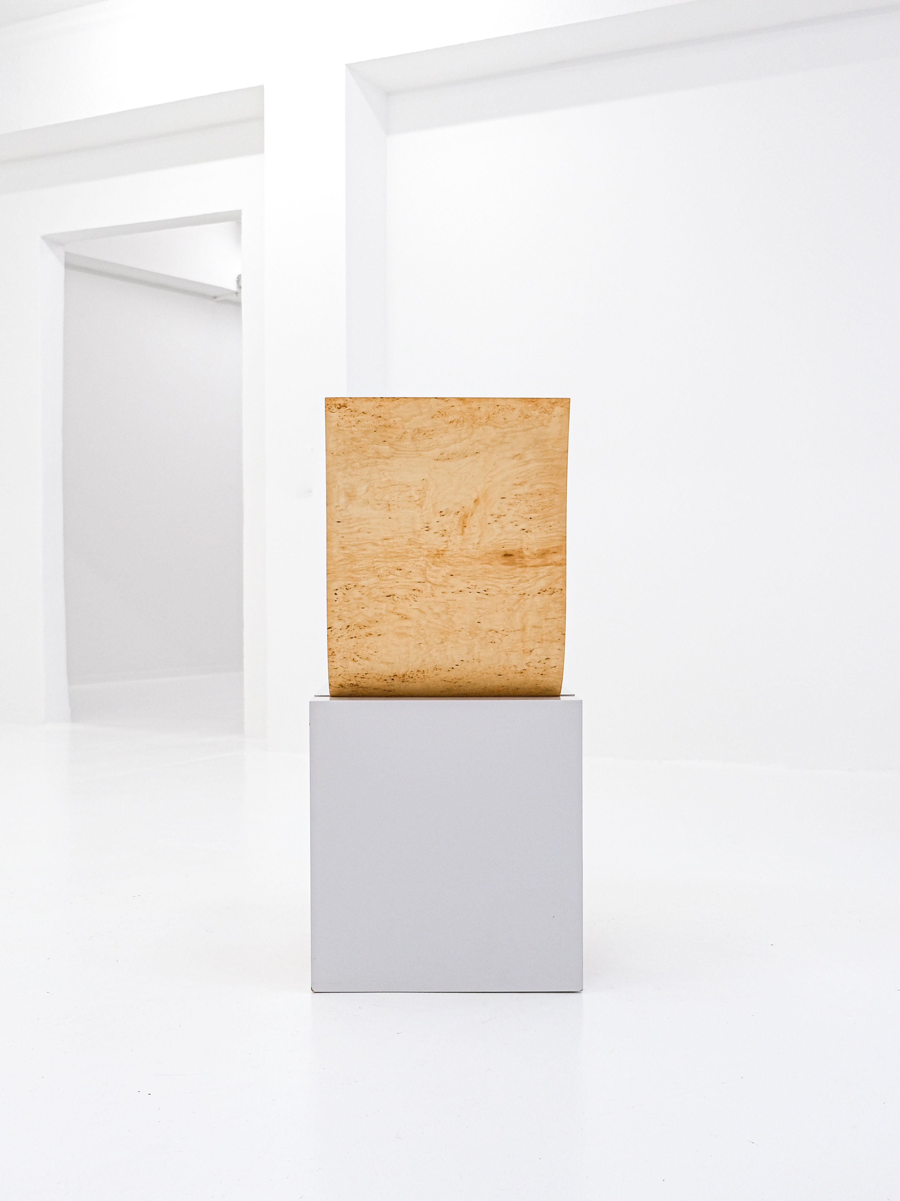 A minimalist wooden sculpture, echoing the Postmodern Leda Chair by Angela Oedekoven for antibeige (1990), adorns a white pedestal in an airy gallery. Its natural wood texture contrasts with the white wall, with an open doorway in the background.