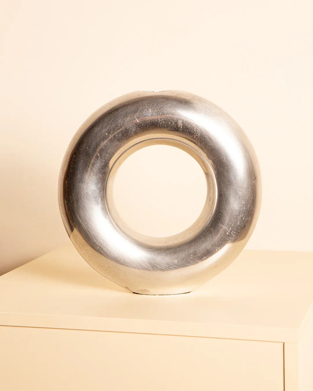 The Treaptyque 80's Aluminum "Circle" Vase, an Italian-style polished metallic donut sculpture, sits on a light beige pedestal against a cream-colored background, beautifully reflecting light with its sleek and smooth surface.