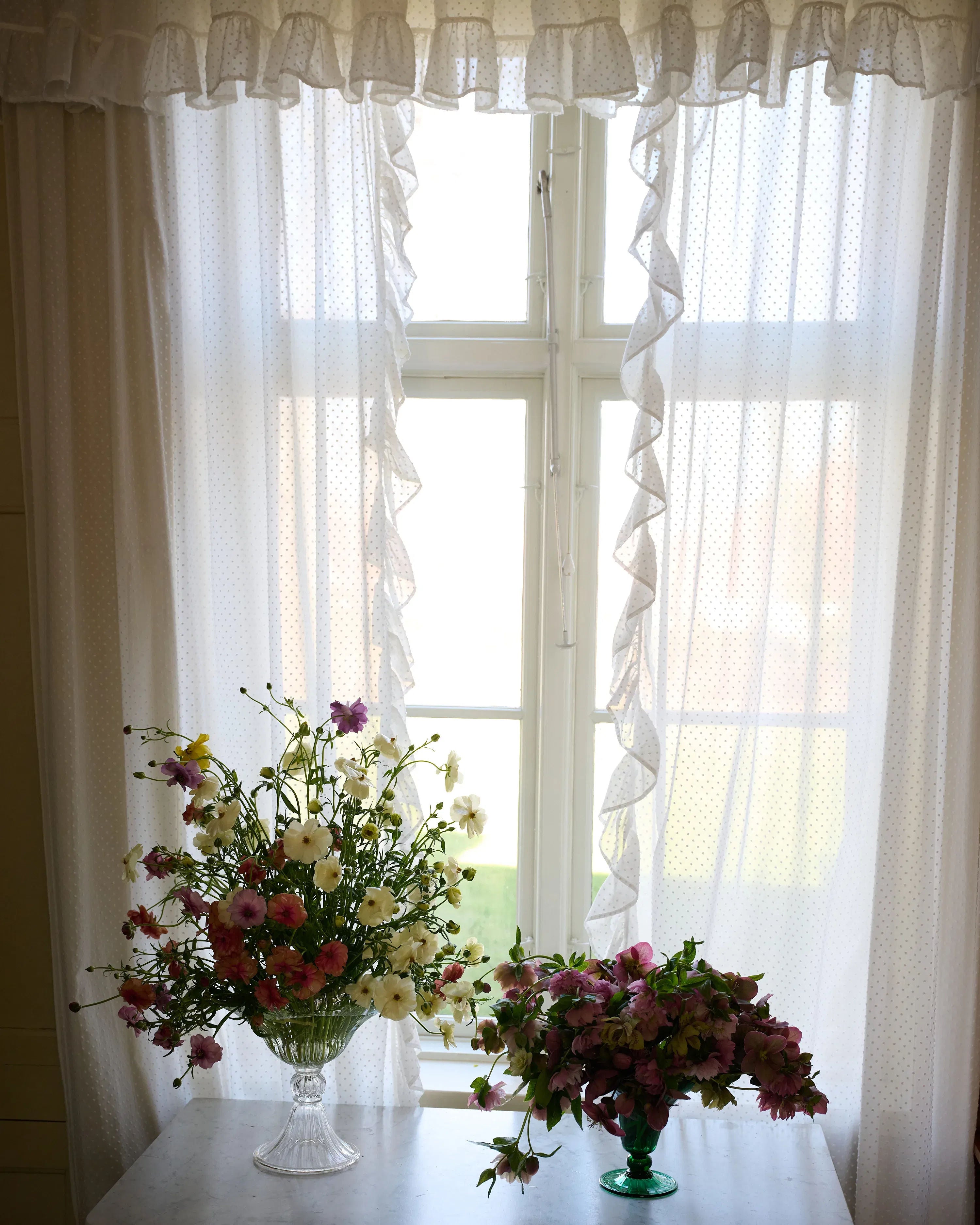 A sunlit window with white, ruffled curtains features a pair of floral arrangements on a table: a clear pedestal vase with a mix of pink, white, and yellow flowers on the left, and an Akua Objects Karen Blixen Vase Small with pink flowers crafted from exquisite Murano craftsmanship on the right.