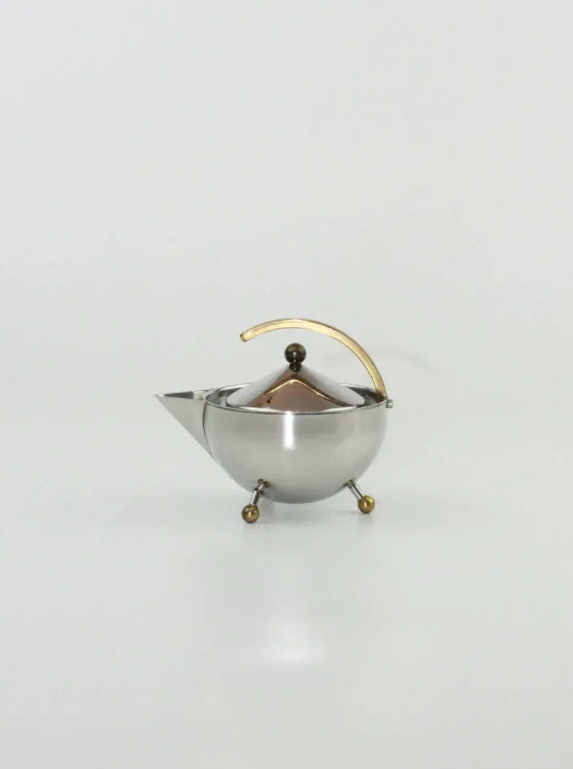 A sleek, modern metal Danish Sugar and Creamer Set by Boga Avante Shop features a spherical body, short spout, and a unique curved gold handle. Crafted from stainless steel and brass, it stands on three small, gold-tipped legs. The matching lid with a small gold knob completes this Boga Avante Shop masterpiece against a plain white background.