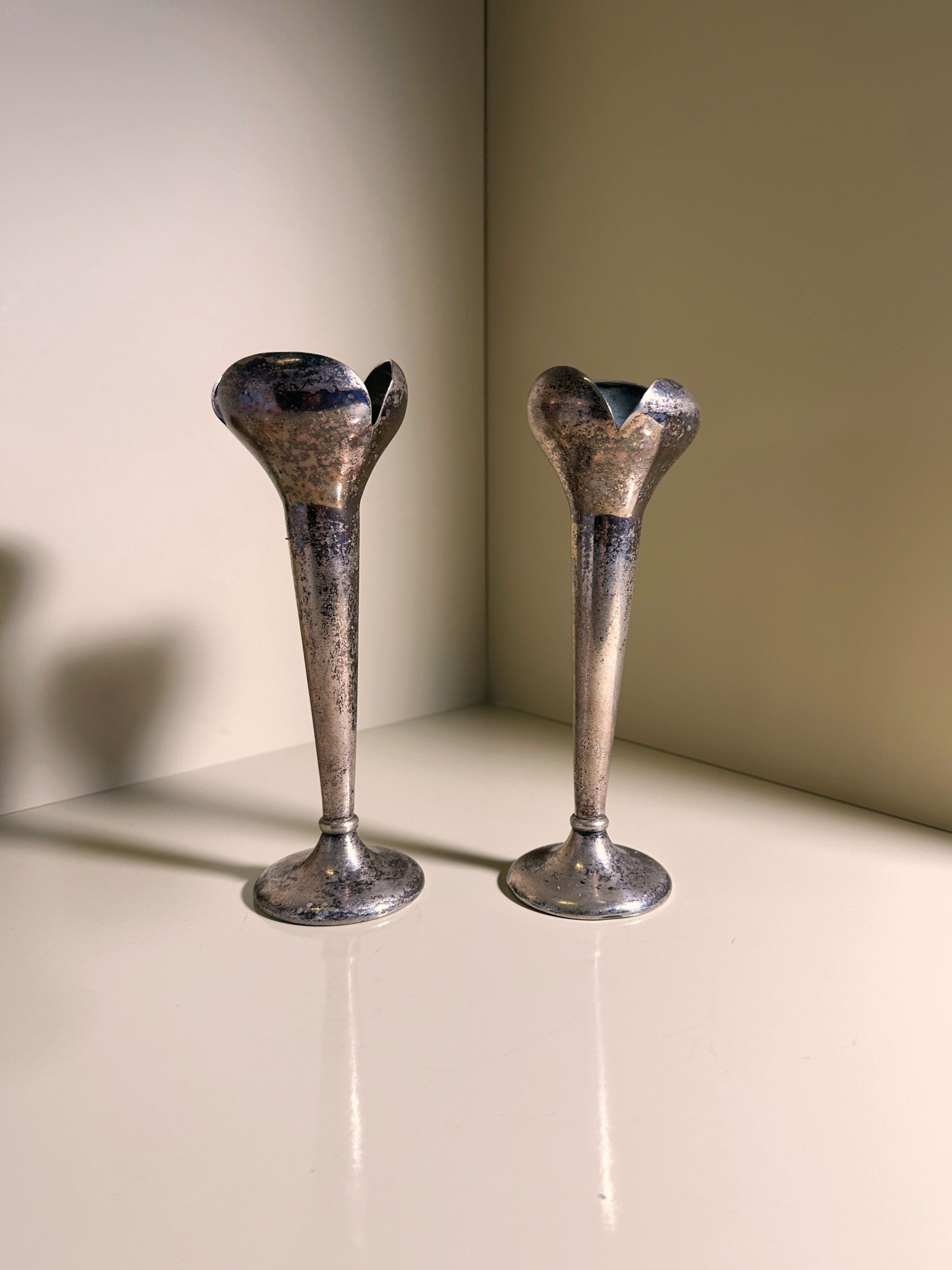 A set of two metal candlesticks by Gio Ponti for Calderoni from the 1930s, provided by Dodo Vintage, stands on a light surface. With their tarnished, antique finish, they cast soft shadows that capture timeless elegance.