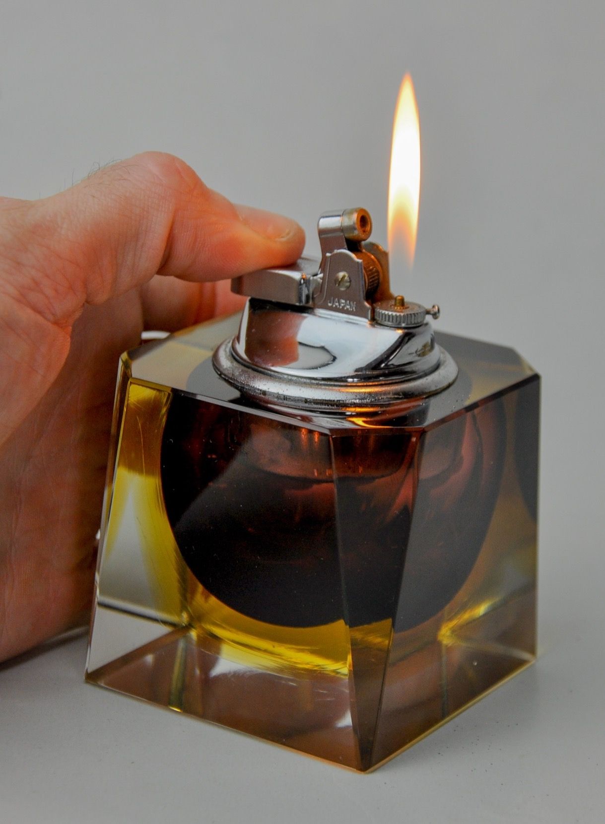 A person's hand holds the Sommerso Lighter by Seguso from Malamar Studio, featuring a vintage square glass design with a metal top. Its small flame flickers against the gray background, and its amber-hued Murano glass body evokes Flavio Poli's masterpieces from 1970s Italy.