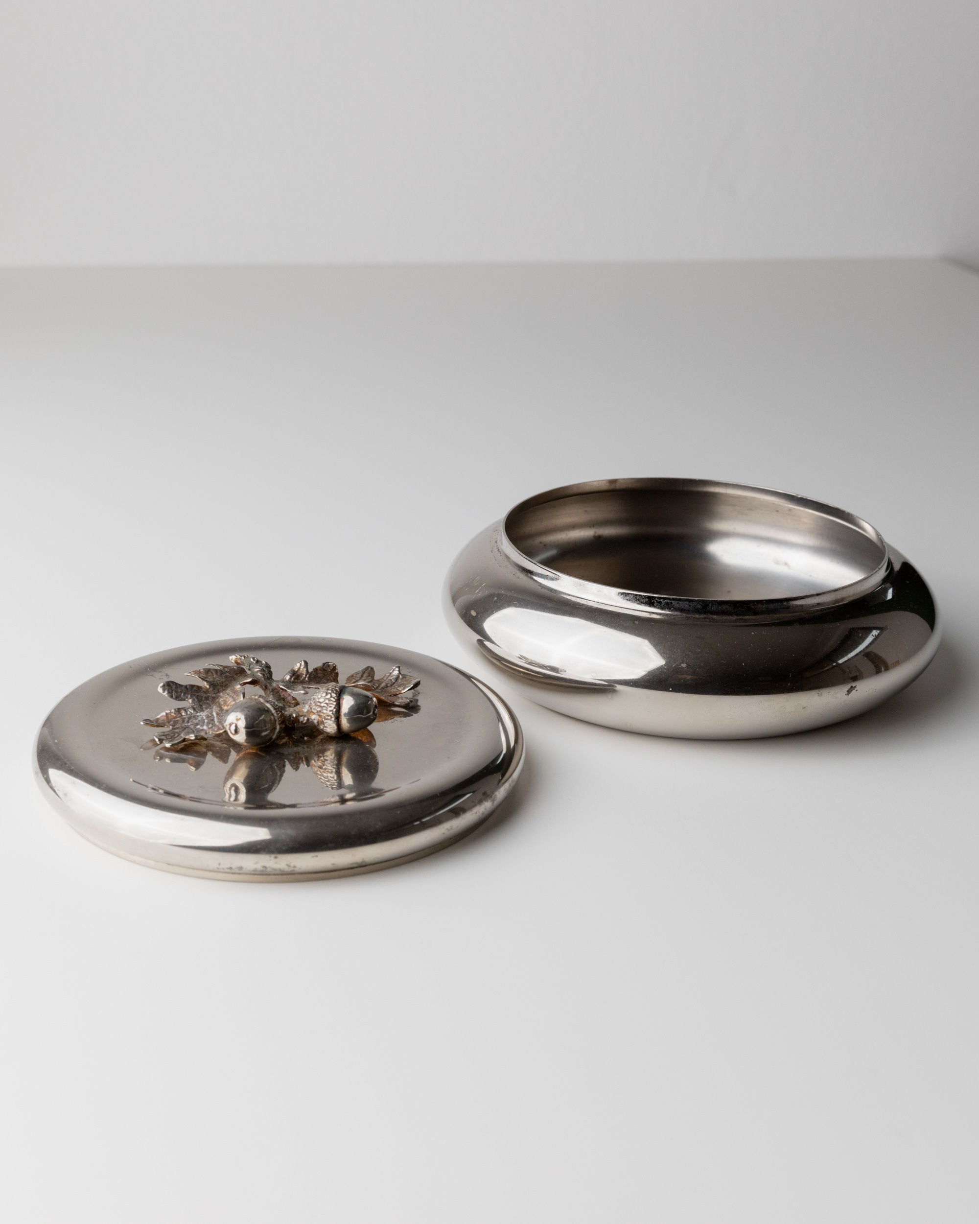 A Silver-plated Box by Bottega Jacobs, Italy 1950s, sits open on a white surface. Its lid showcases ornate silver leaf designs and embedded stones, featuring an acorn handle. The box's smooth, polished finish reflects light like a vintage tin.
