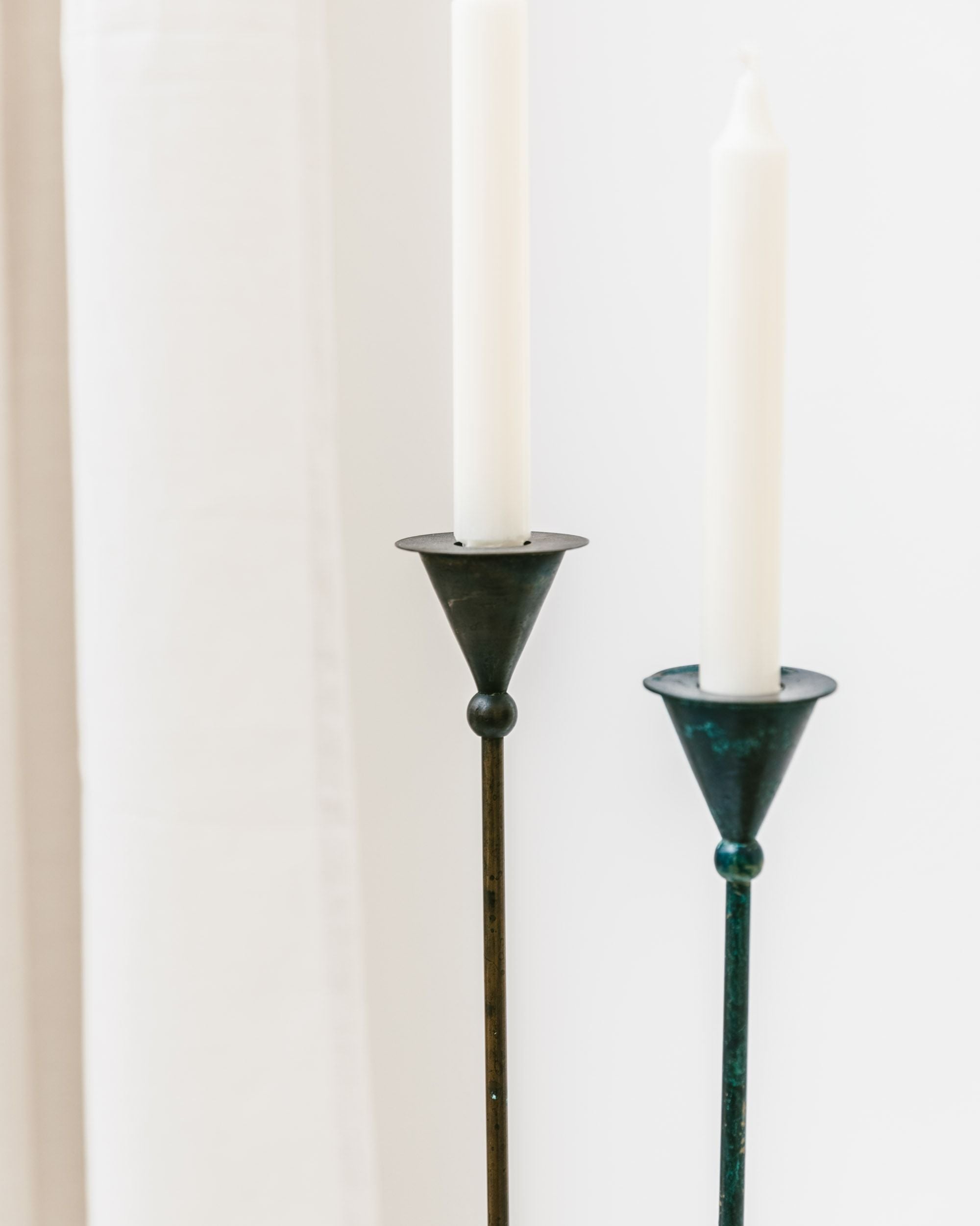 The Set of 2 Candleholders by Bottega Jacobs, hailing from the Postmodern 1980s, features two tall and thin candlesticks in a vintage style holding unlit white candles. The left candleholder displays a slightly taller conical form, gracefully complementing the right. A softly blurred background highlights their minimalist and elegant appearance.