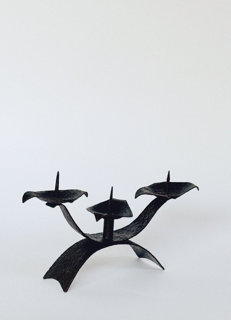 The Huerto 3 Arm Metal Candleholder boasts a mid-century artisan design with three wrought iron branches featuring pointed tips and elegant curved, leaf-like platforms for candles, set against a plain white background.