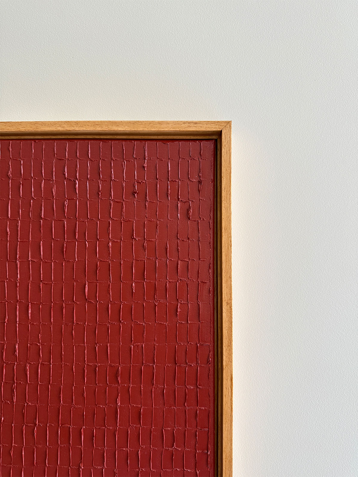 A framed artwork with a textured red surface, featuring a grid-like pattern reminiscent of Korean Dansaekhwa, is hanging on a plain white wall. The wooden brown frame borders the piece cleanly, adding depth and interest to the minimalist composition. The painting titled Indian Red - Oil on canvas by Roi Elmaliah adds an elegant touch to the overall aesthetic.