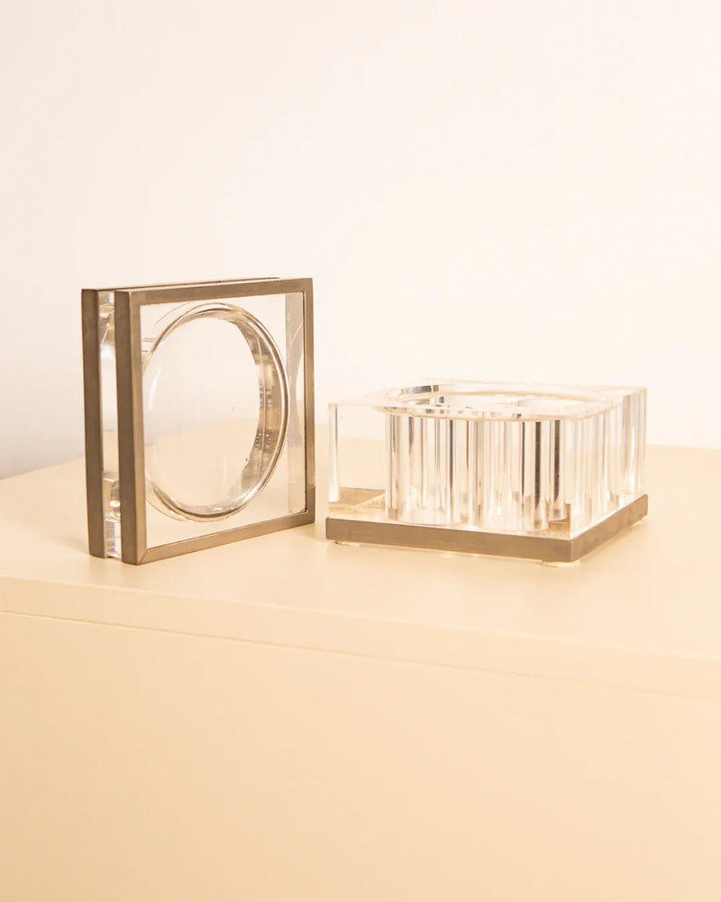 The Metal and Plexiglas box by Soldi & Scati from the 70s, designed by Treaptyque, features two transparent geometric objects with metallic edges resting on a beige surface. A cube and a rectangular prism exemplify sleek, modern Italian design with their clear sections and reflective materials that elevate their minimalist style.