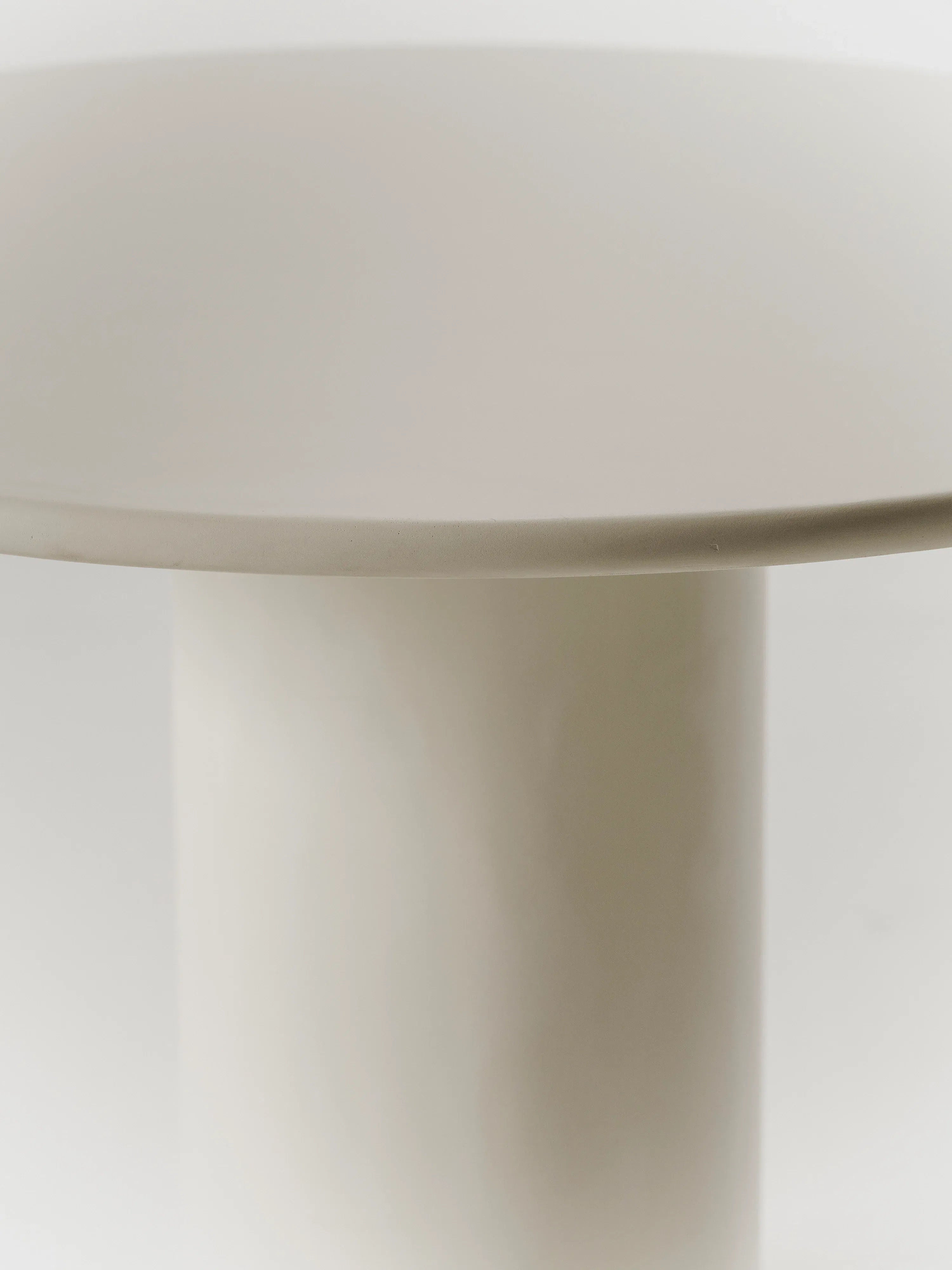 Close-up of MarlotBaus' Gio Dining Table: a minimalistic, round, flat beige tabletop with a cylindrical base against a light background, highlighting the smooth surface and modern design of this microcement piece.