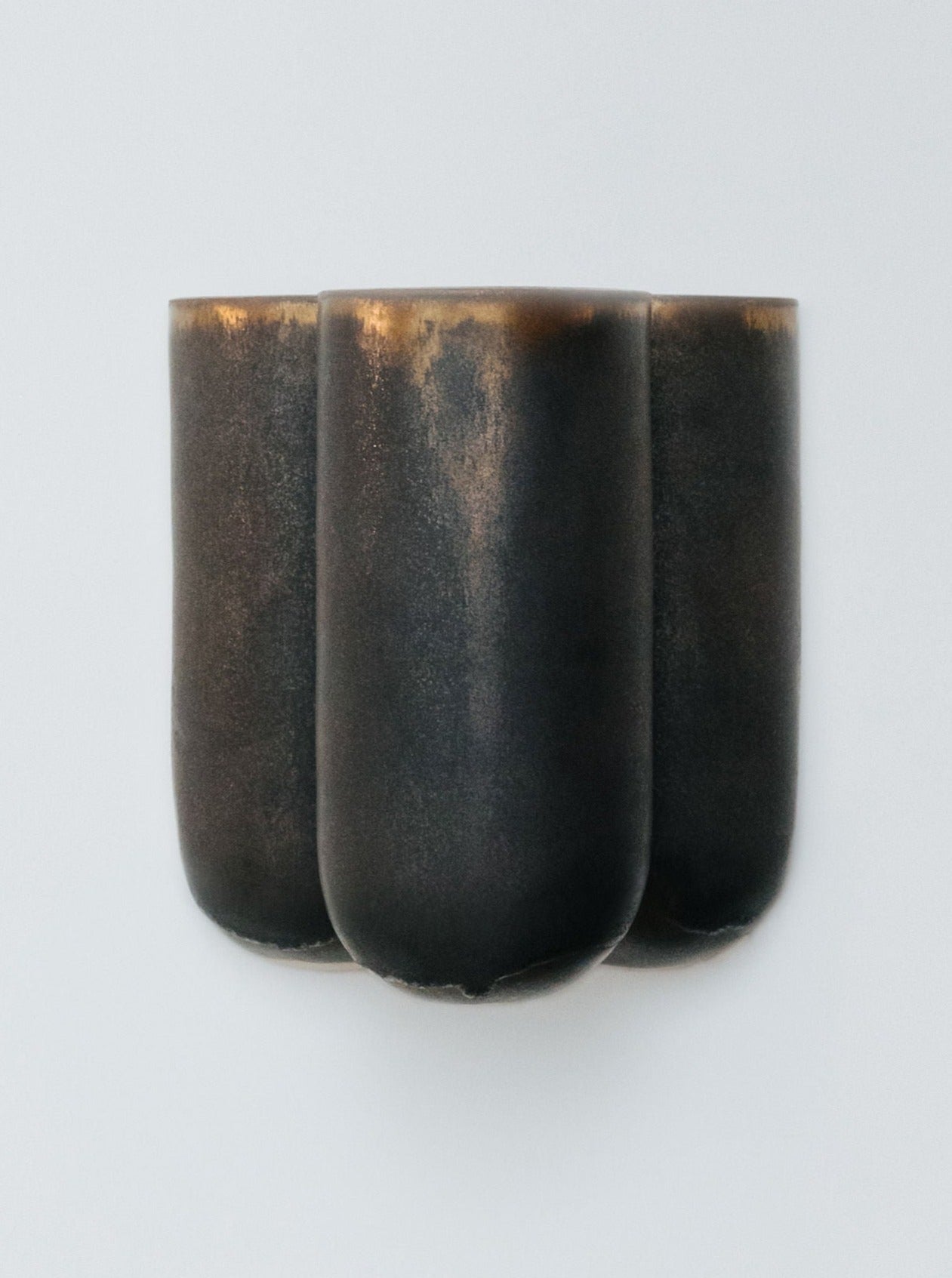 The Wall light "MOOR" by Lisa Allegra is a minimalist piece featuring a dark bronze design with sculptural elegance. It showcases three cylindrical forms seamlessly connected, displaying a smooth texture adorned with glaze effects that create subtle color variations against a plain white backdrop, reminiscent of the refined style found in MOOR wall lights.