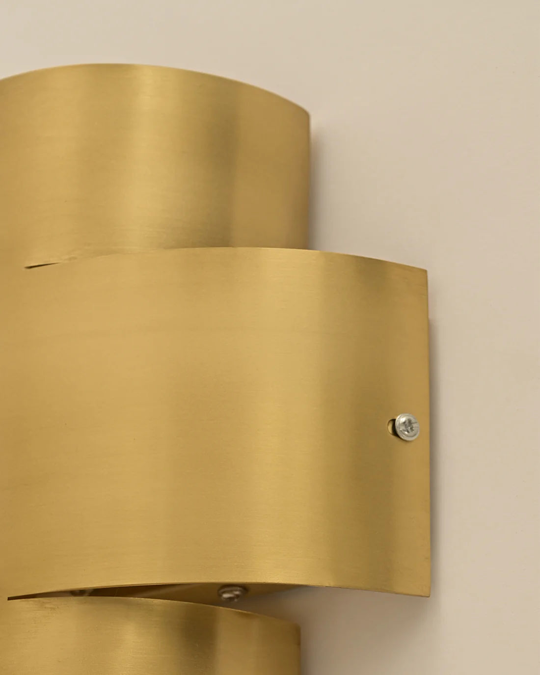 Close-up of the Aura Wall Sconce by LMNOH, featuring a brass structure with a smooth gold finish. This fixture embodies modern art with its sleek, minimalist design and visible screws securing sections against a light wall—ideal for contemporary interiors.