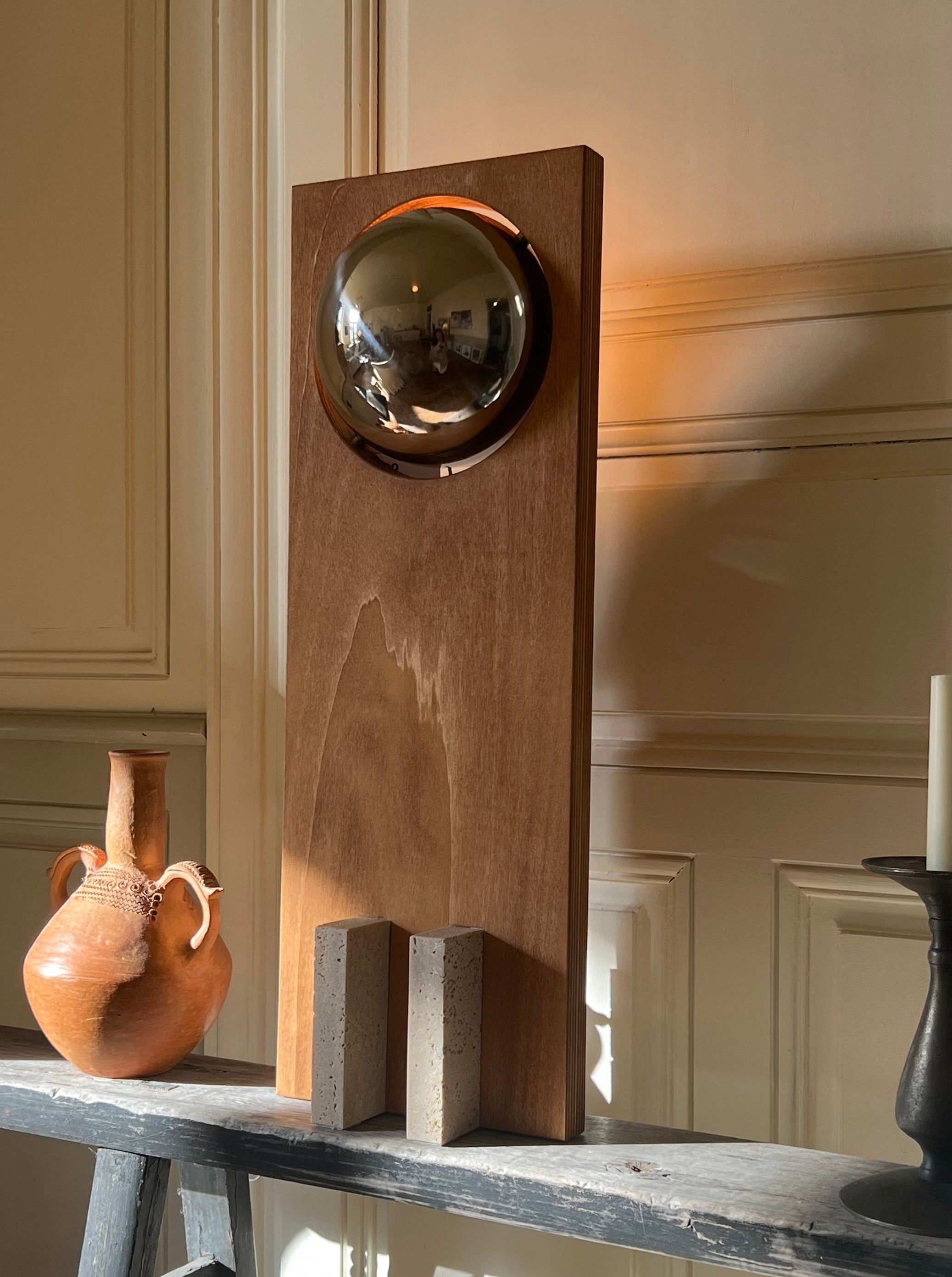 A modern art installation featuring a Galileo 2.3 reflective spherical object mounted on a tall wooden panel. Beside it, on a gray shelf, sits a rustic clay pot and a simple candlestick. Soft lighting from a Edoardo Lietti Studio.