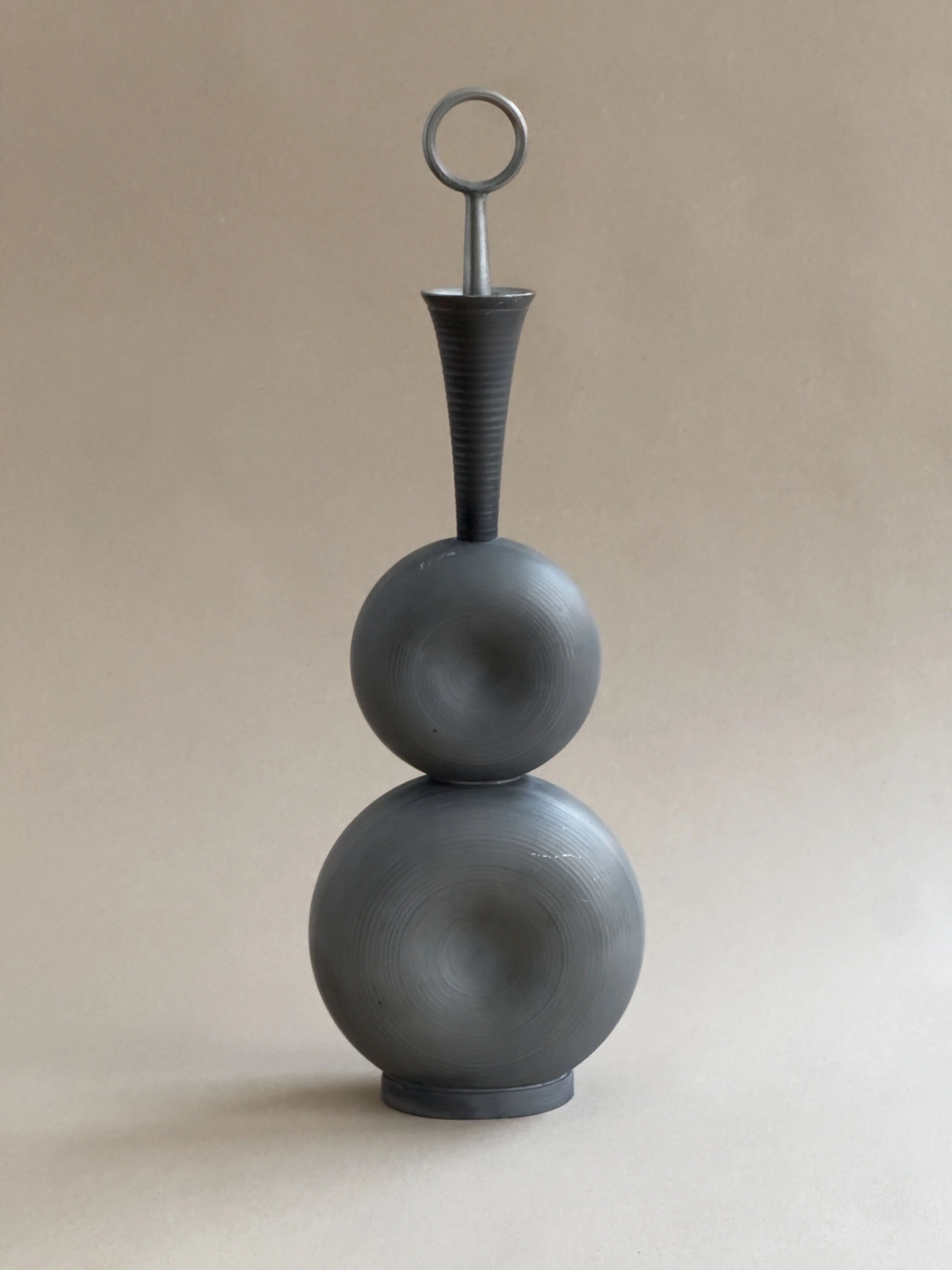 The Galerie Storm Modernist Pewter Decanter, featuring a gray design with two spherical tiers and a tall neck topped by a circular handle, echoes Gunnar Havstad's artistic flair with concentric patterns and sits against a neutral background.