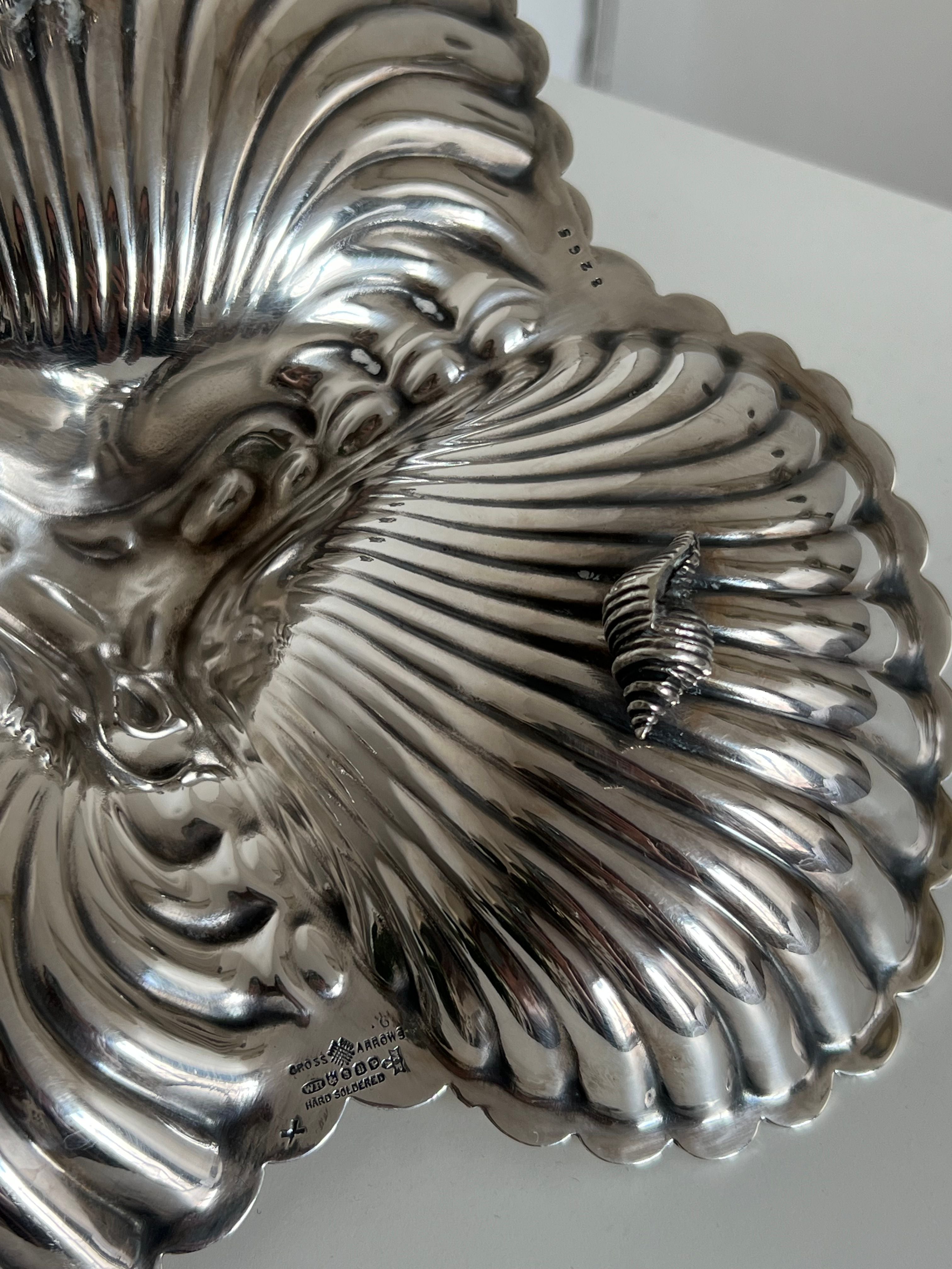 A close-up photo of a shiny, decorative Triple Shell Silver Shell Serving Dish by Manushi with a scalloped, ridged design, evoking the appearance of seashells. The dish features intricate details and a small, ornate handle shaped like a screw on the right side. The background is plain white.