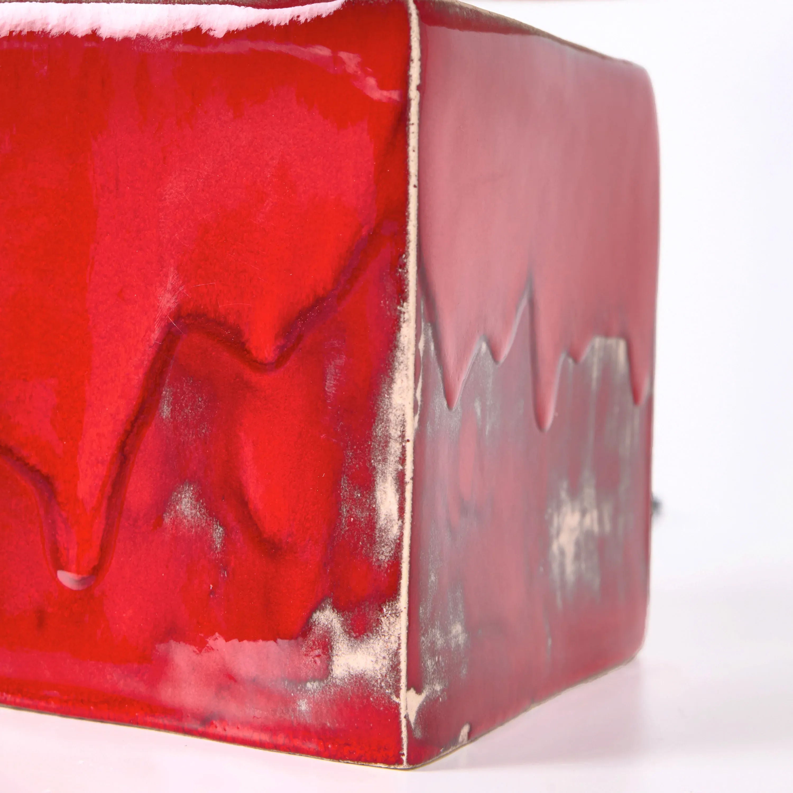 A close-up of Project 213A’s Arouca Table Light Red shows its glossy red ceramic cube base with smooth, flowing red and white glaze patterns on two sides, resembling melted paint. The background is plain white.