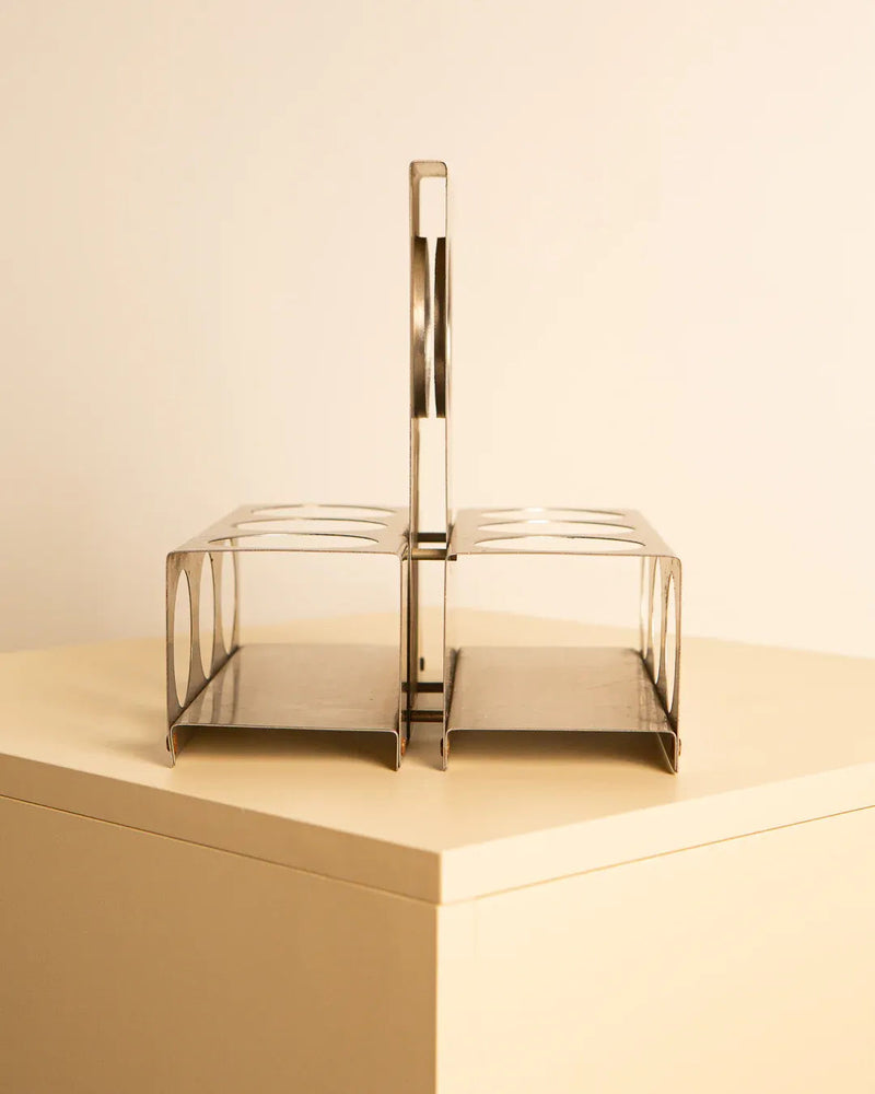 A Brushed Steel Wine Rack by François Monnet 70's, available from Treaptyque, is placed on a beige-colored corner table. The rack, reminiscent of a brushed steel wine rack, has multiple slots for holding slices of toast upright and a handle in the center for easy carrying. The design is minimalist and modern.