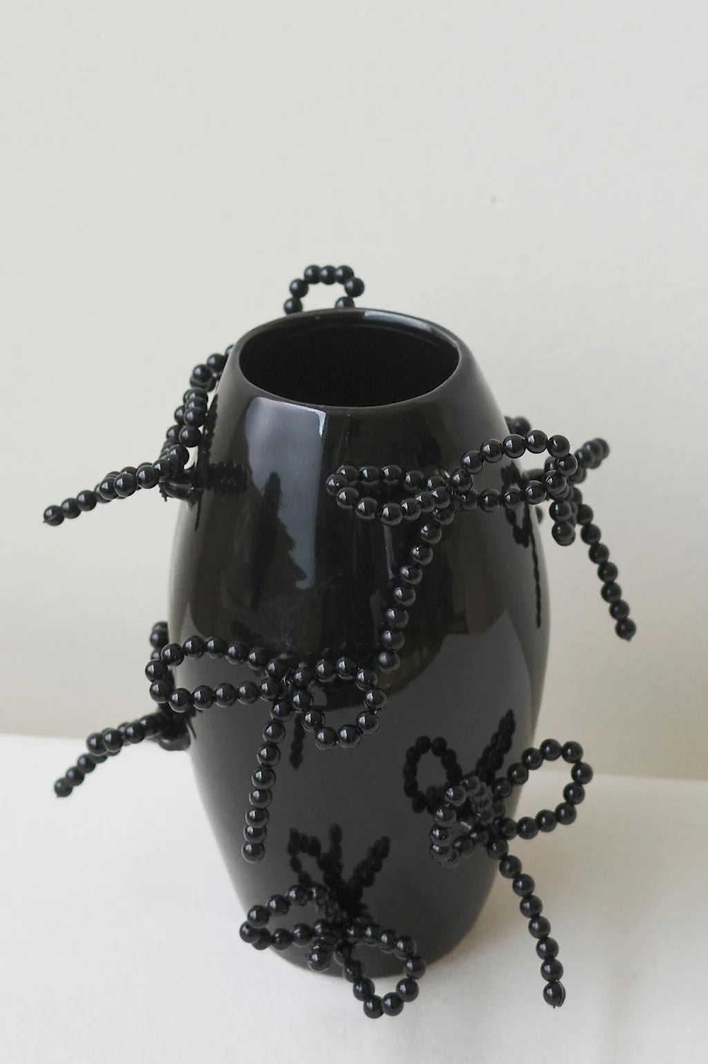 The Pearly Pearl - Small Vase in Gloss Black by Completedworks, with its sculptural form and black beaded bows, stands elegantly on a white plane against a pale backdrop.