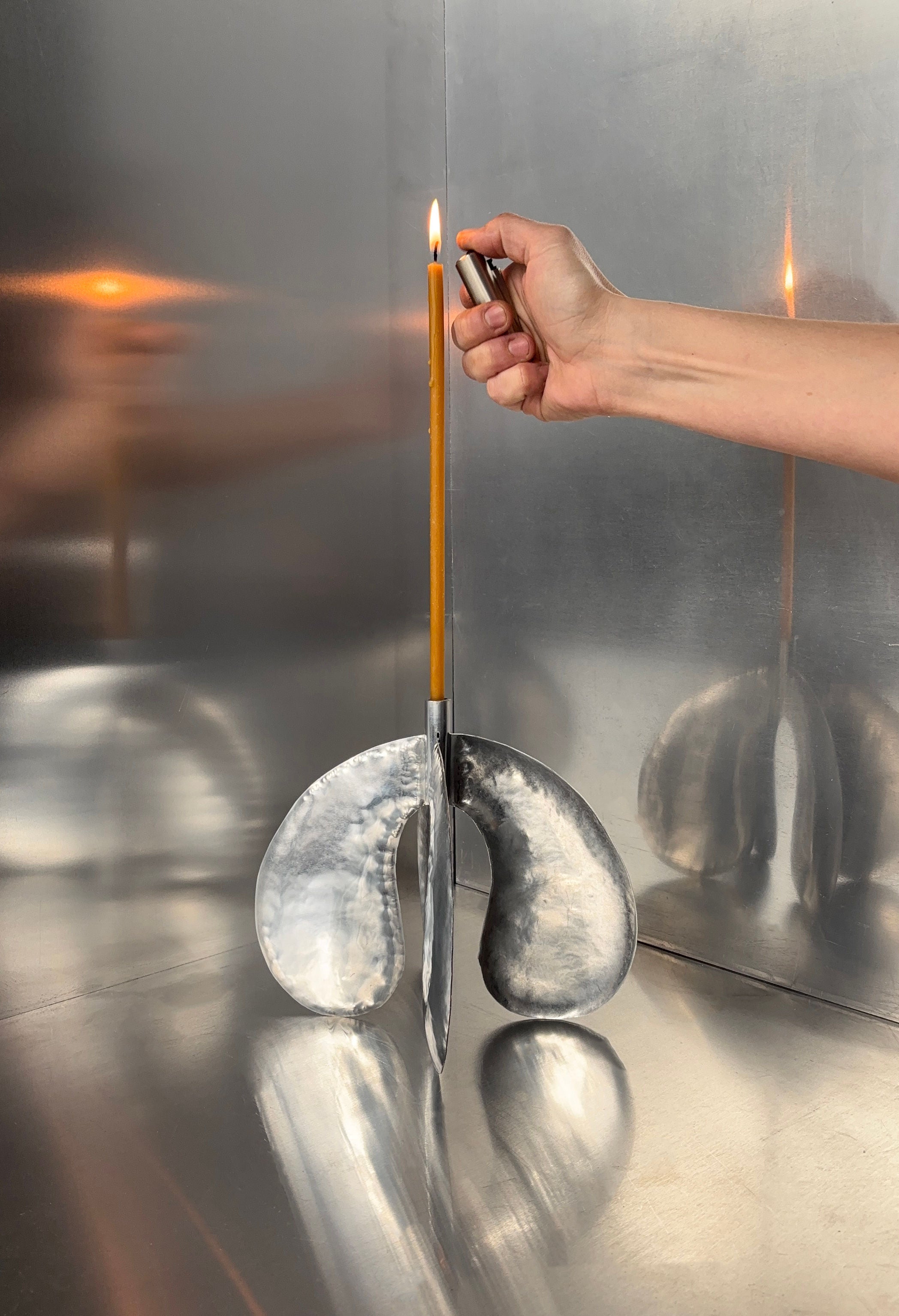 A hand lights a thin brown beeswax candle held in the Bregje Sliepenbeek's Phaedra Large candle holder. The modern, abstract silver design beautifully reflects its flickering flame.