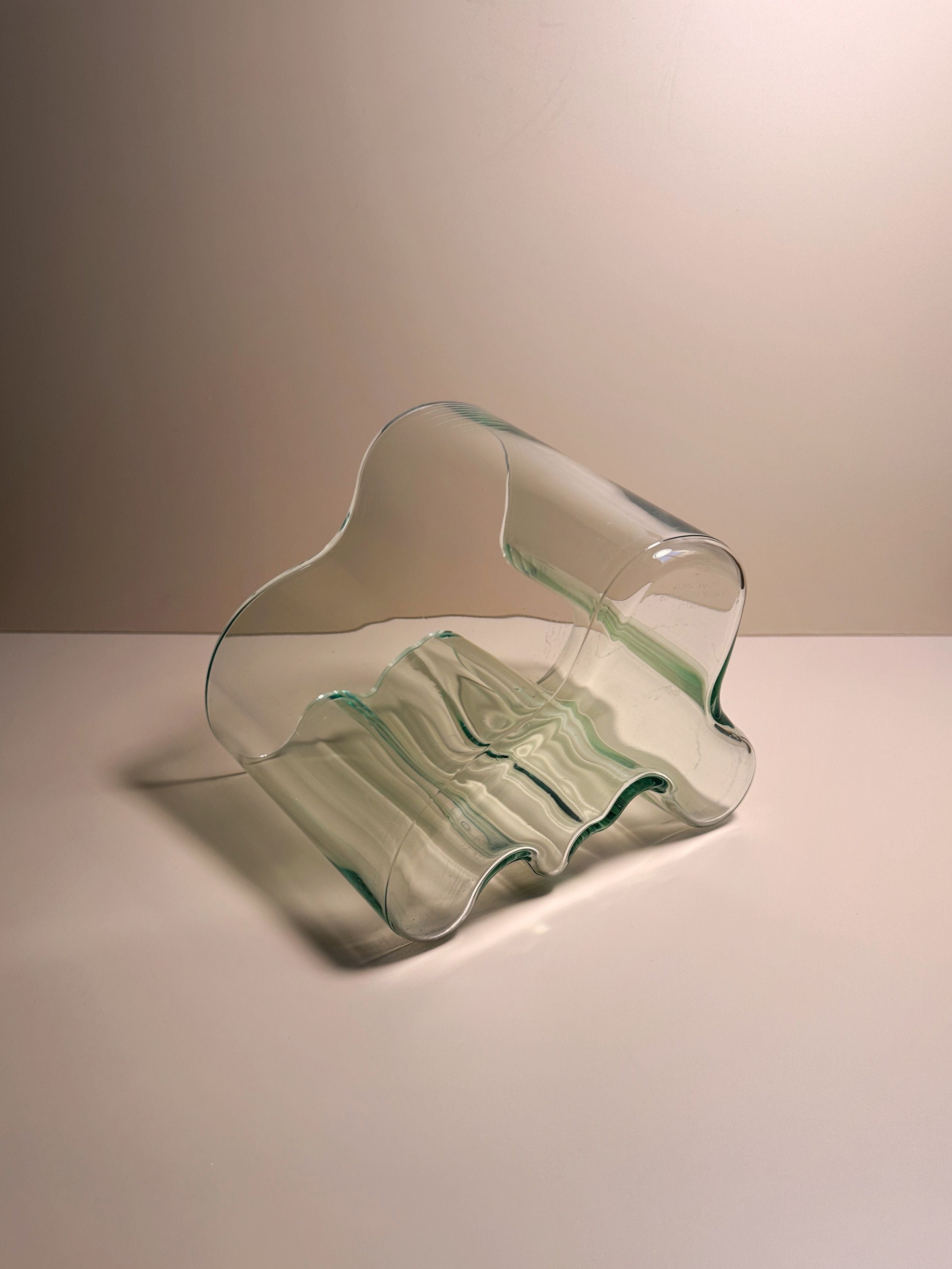 A transparent, wavy glass sculpture with gentle curves, reminiscent of the Limited Edition Aalto Vase by Dodo Vintage, is placed on a smooth, light-colored surface. The delicate design captures and reflects light, creating subtle patterns and shadows. The softly lit background enhances the elegance of this prized collector's item.