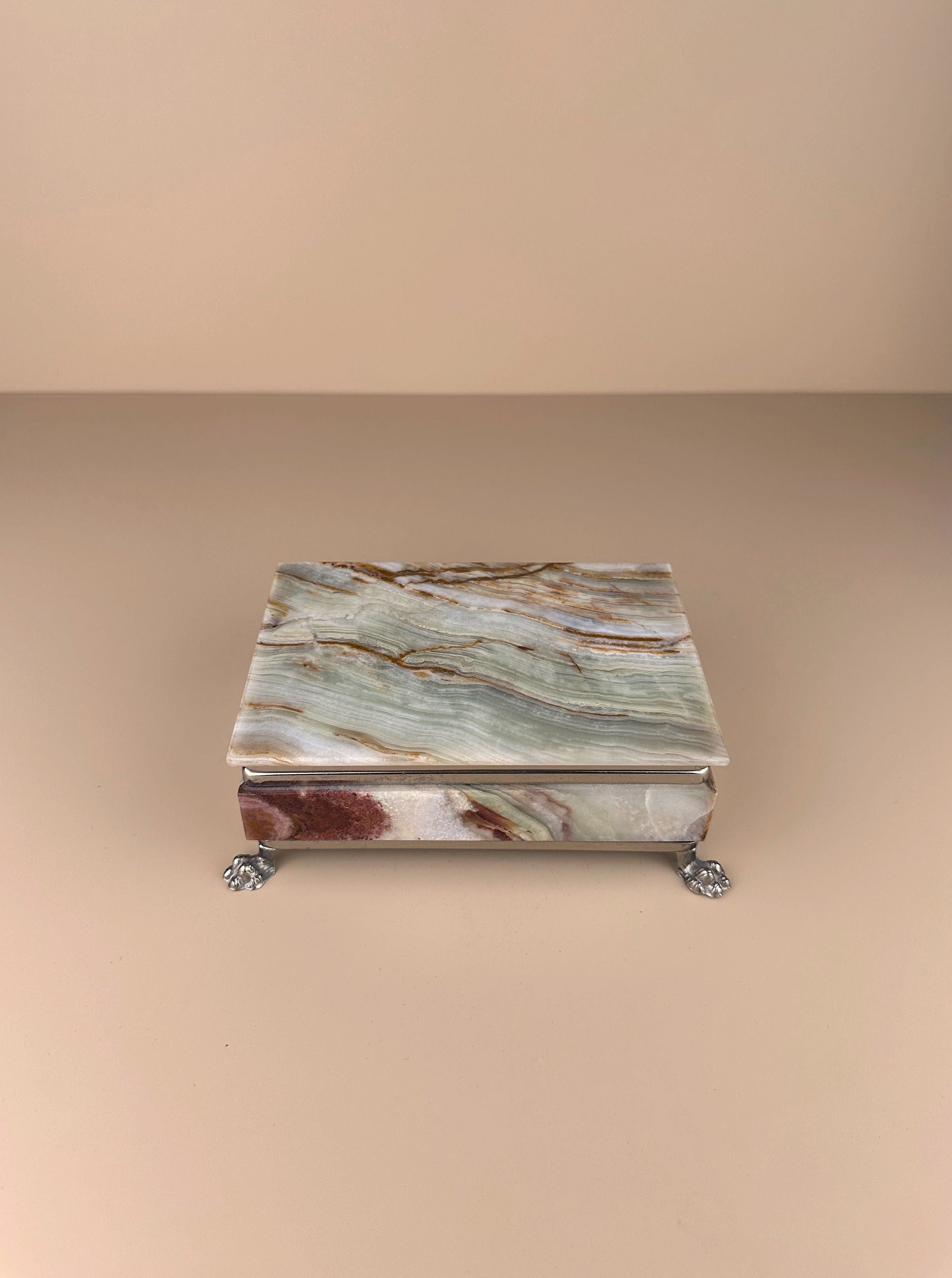 Image of a rectangular, marbled stone box with earthy tones of green, brown, and red. Resembling a vintage Italian jewellery box, it rests on four small, intricately designed metal feet, giving it a slightly elevated appearance. The neutral beige background complements the 1970s Italian onyx jewellery box by Dodo Vintage's natural hues.