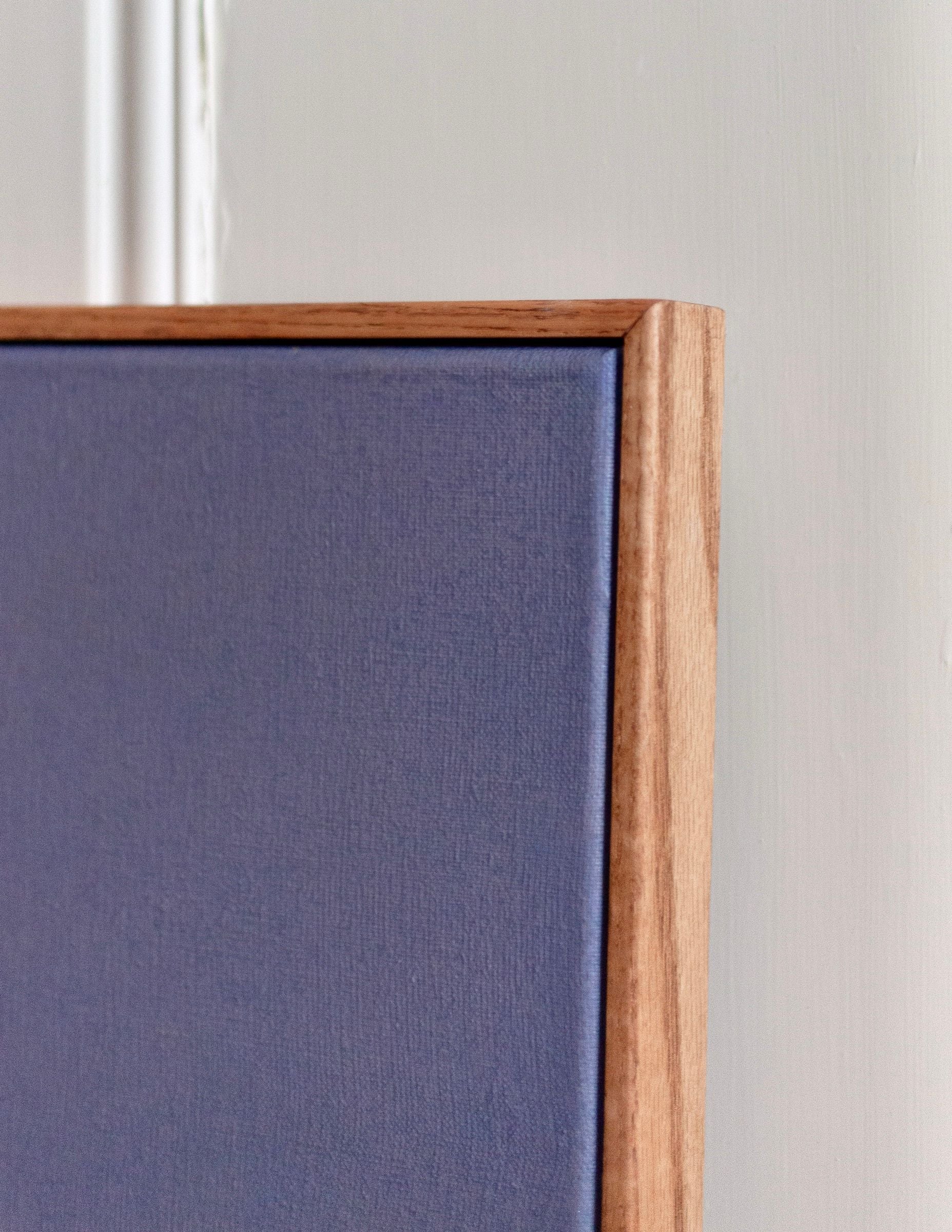 A close-up of 'Seabeds #2' Painting by Rebecca von Matérn showcases an oak frame and a blue fabric panel. The panel contrasts gently against a light wall, embodying the clean lines of Stockholm design, with the wood's warm tone highlighting material differences.