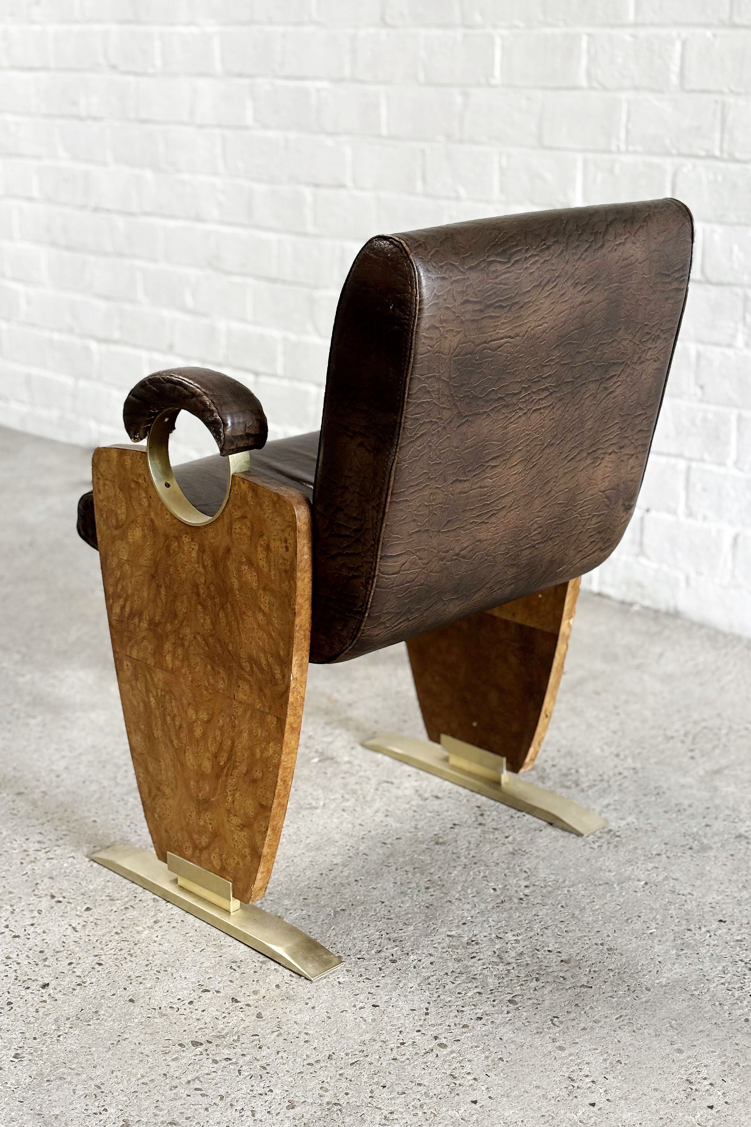 The Introverso Art-Deco Style Side Chair, from the 1970s, features rich skai leather upholstery and a dark brown cushion. It is complemented by burl walnut wood armrests, with a unique triangular base design and metal accents on the feet, set against a white brick wall background.