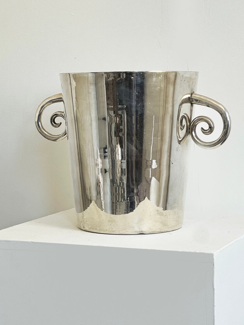 The Spiral Handle Ice Bucket by Porter Studio, featuring two ornate curved handles, sits on a white pedestal against a plain white background.
