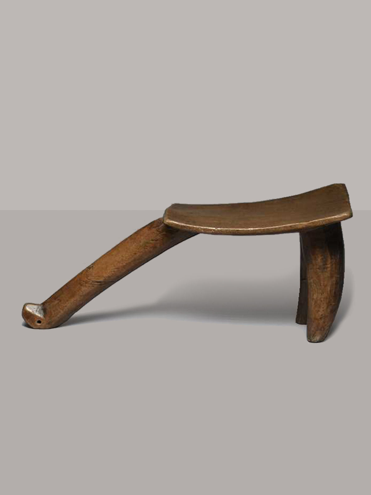 The Lobi Tripod Stool 1 by RELIC LONDON is a traditional wooden headrest with three legs, resembling an African tripod wooden stool from Burkina Faso. It features a curved, flat top surface for resting the head. The minimalist design and worn finish highlight its vintage condition, indicating its age and use.