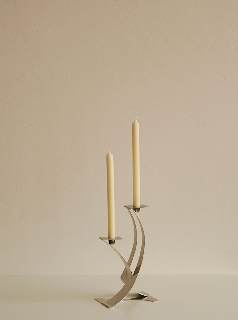 A Les Objets Amsterdam Art Deco Candleholder with two beige candles in a modern, geometric design stands on a neutral surface against a plain, light-colored wall. The unique candelabra has a sleek, double-arc shape, giving it an elegant and contemporary look.