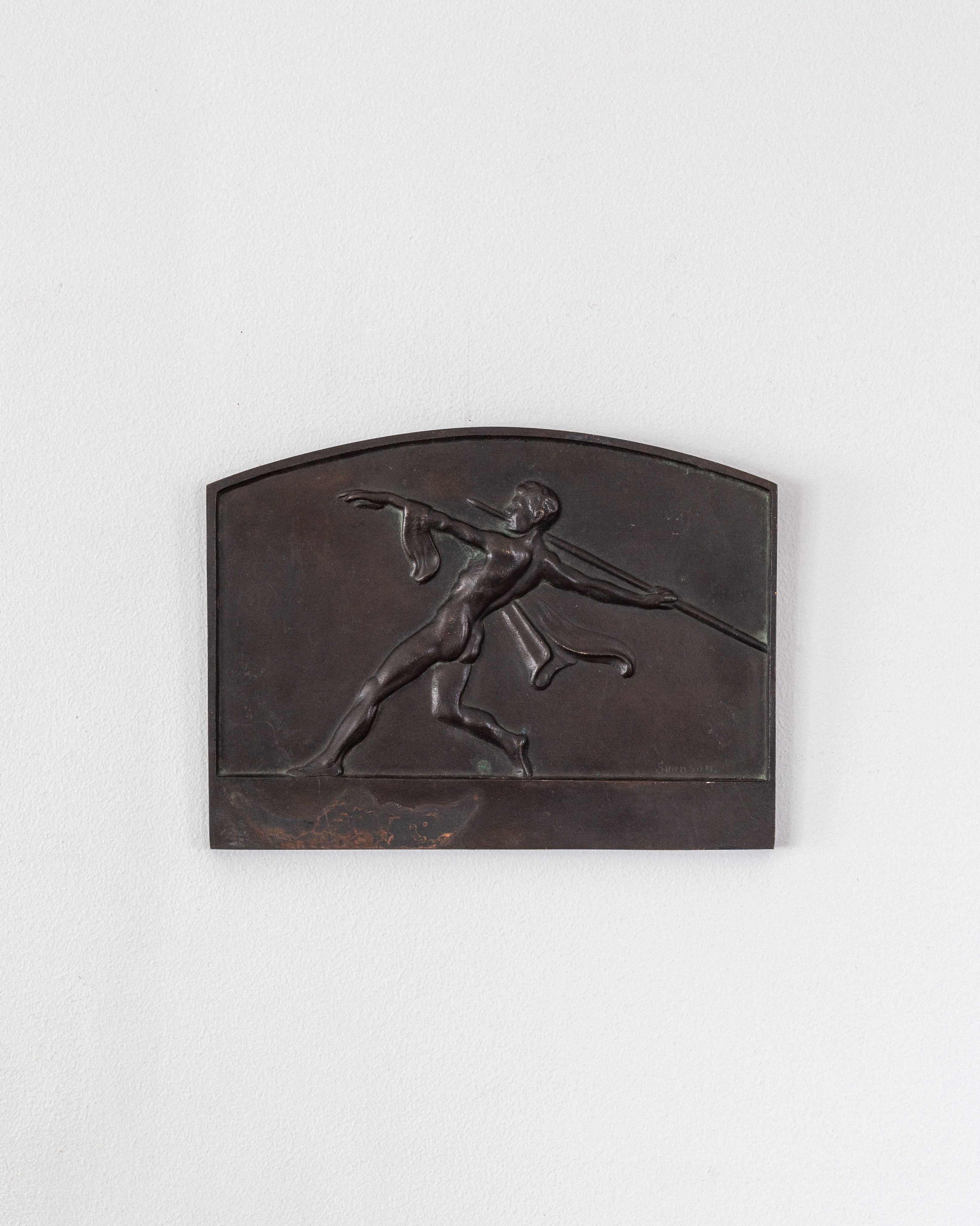 The Spigel "Art Deco Bronze Wall Relief" features a nude figure in motion with a javelin, set against an arched rectangular background. This elegant wall plaque blends classical imagery and modern elegance on a white wall, exuding Art Deco flair.