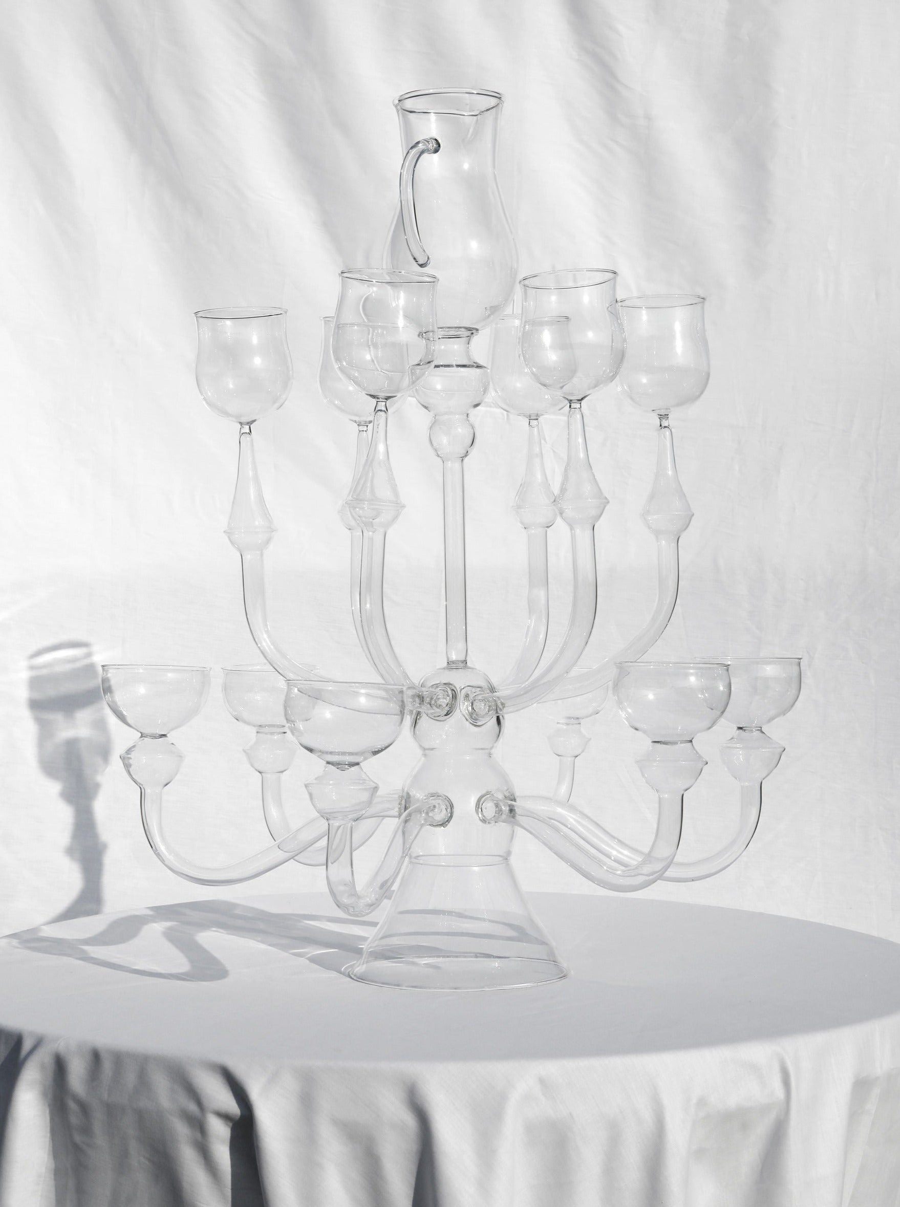 The "Dinner's Ready" Glass Sculpture by Ylaria Pavone is showcased on a round, white table. This large and intricately designed piece features multiple branches holding clear glass goblets and a central glass pitcher, set against a wrinkled white fabric backdrop to create a unique dining experience.