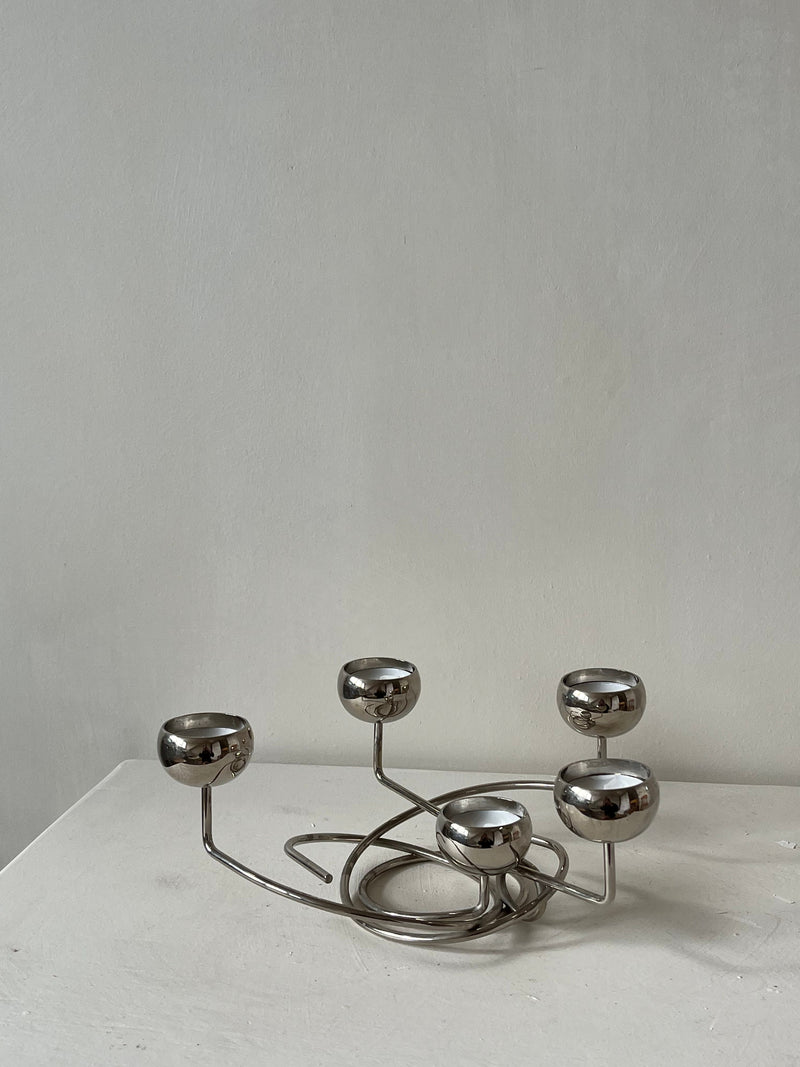 Introducing the Chrome Candleholder by septembre studios: a modern, sculptural piece featuring five rounded holders on an elegantly intertwined base. Its unique angles stand out beautifully against a plain gray background on a white surface.
