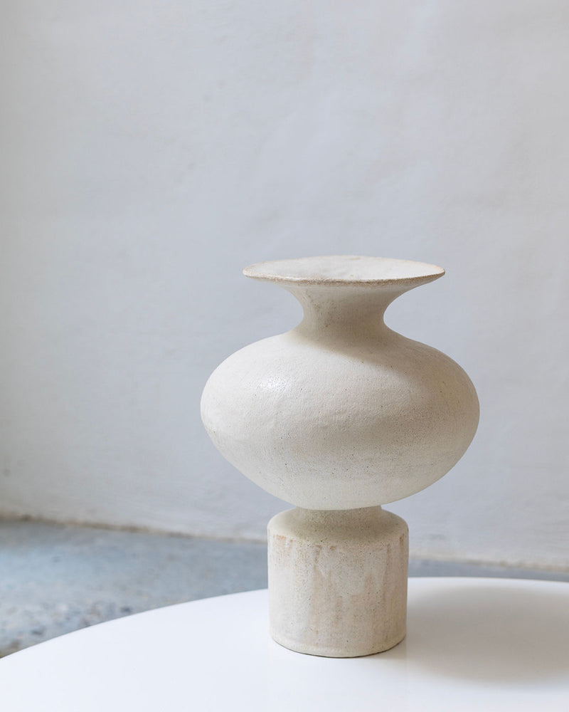 The Isolated n.11 Perla Vase by Canoa Lab, a cream-toned textured ceramic with a wide, rounded body and flared rim, stands on a cylindrical base atop a white surface against a minimalist light gray background.