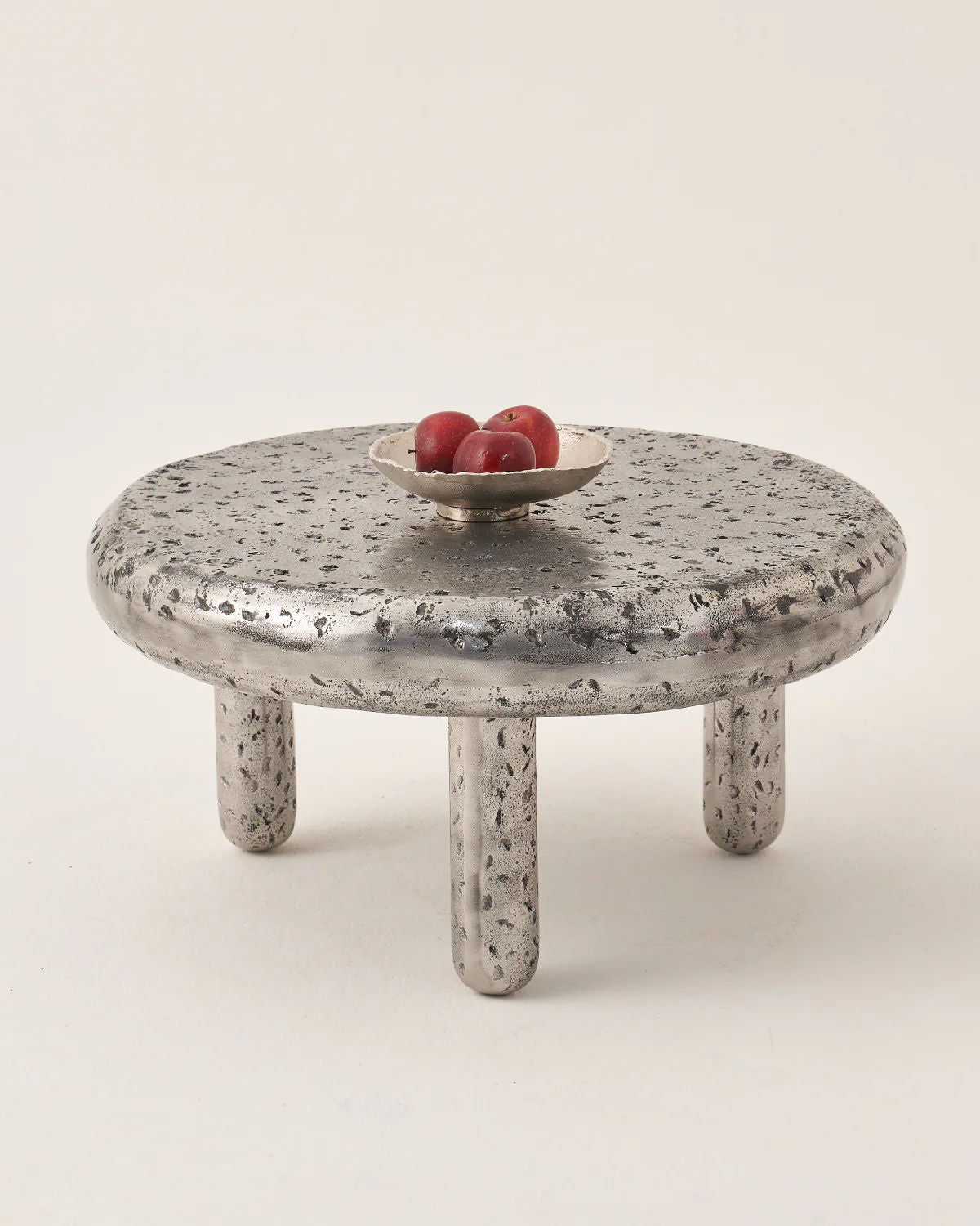 LMNOH's "The Crater - Set of 2" features a round, textured metal table with three legs as a celestial centerpiece. A small bowl on top holds three red apples, set against a simple off-white background for a modern interior vibe.