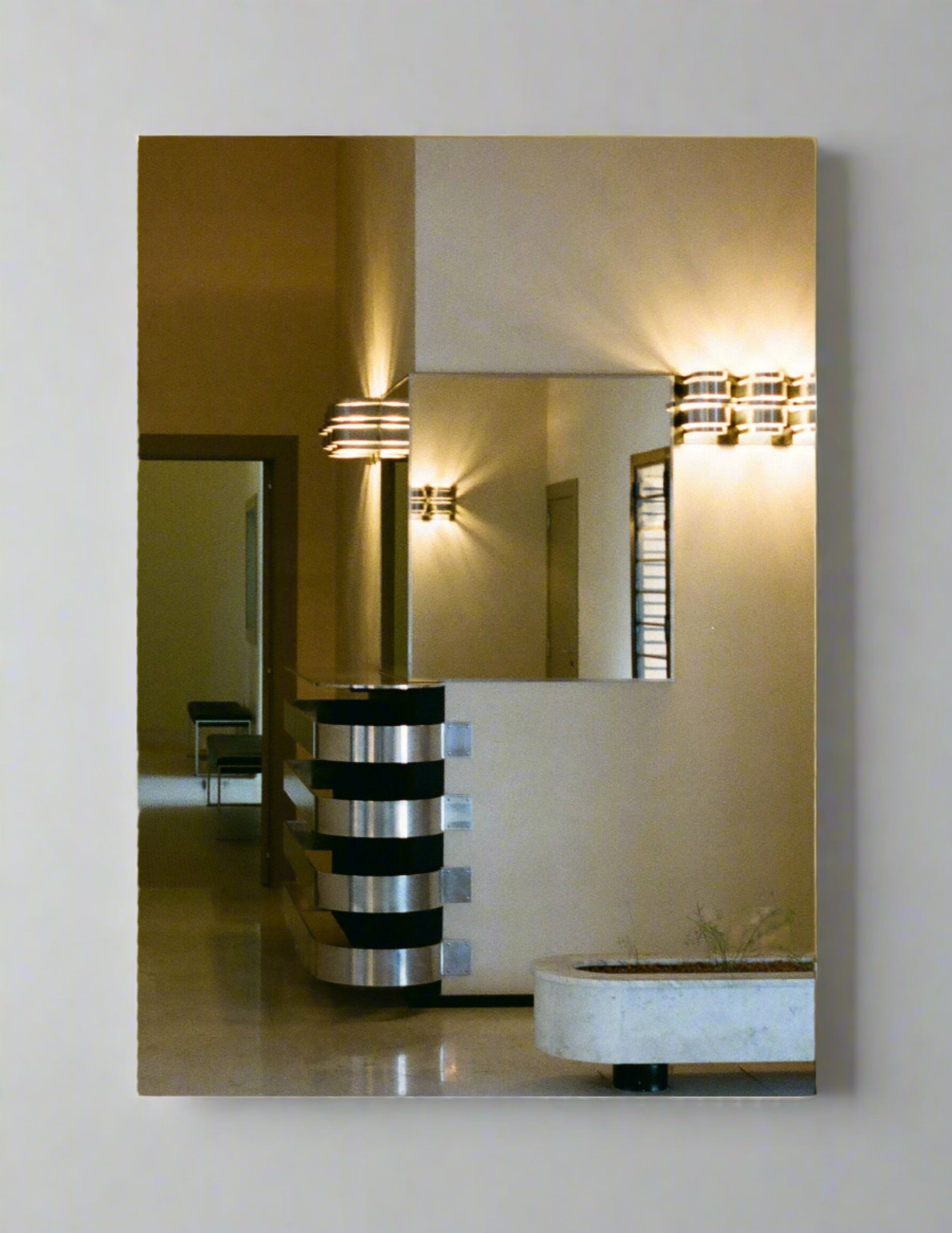 A modern, minimalist interior showcasing a hallway with a mirror reflecting the adjacent wall and part of the corridor. The room features sleek black and silver wall sconces providing warm light, a staircase with horizontal balustrades, and a planter in the corner—captured elegantly in Grace Rose Watts' Villa Cavrois, Entrance - Print Photography.