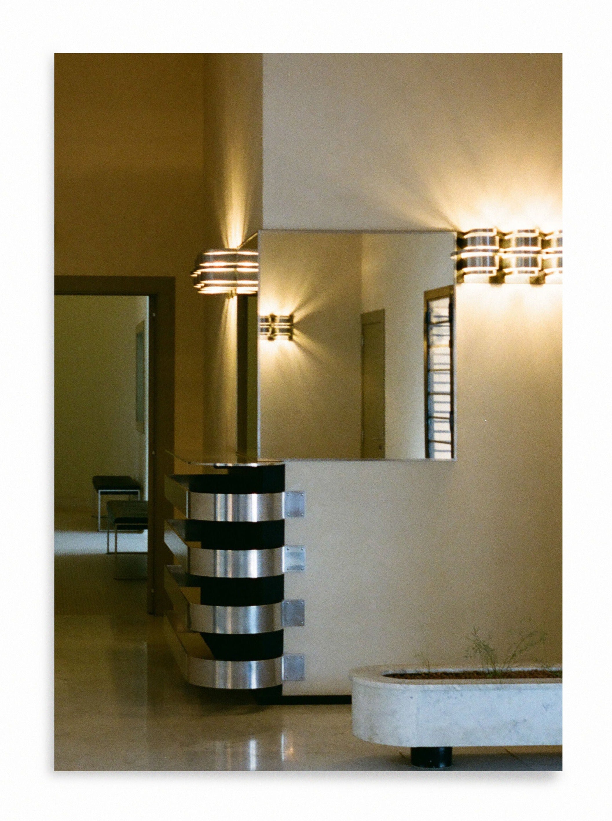 A minimalist interior featuring a sleek marble counter with chrome accents, illuminated by stylish, art-deco wall sconces. A hallway in the background leads to chairs against a wall, where Grace Rose Watts' Villa Cavrois, Entrance - Print Photography creates a clean and modern aesthetic.